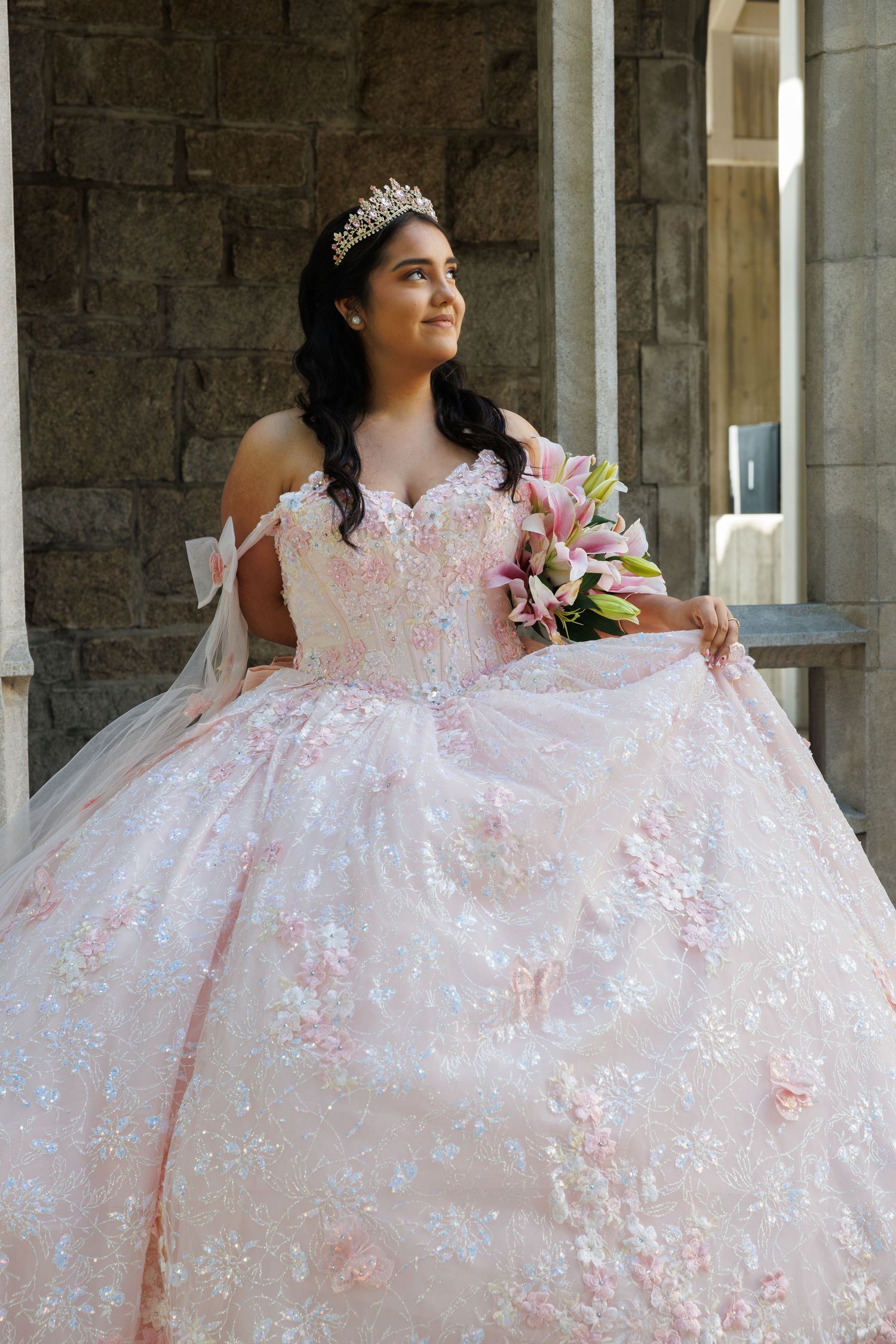 Young woman in pink floral ball gown with tiara holds lily bouquet at quinceañera celebration.