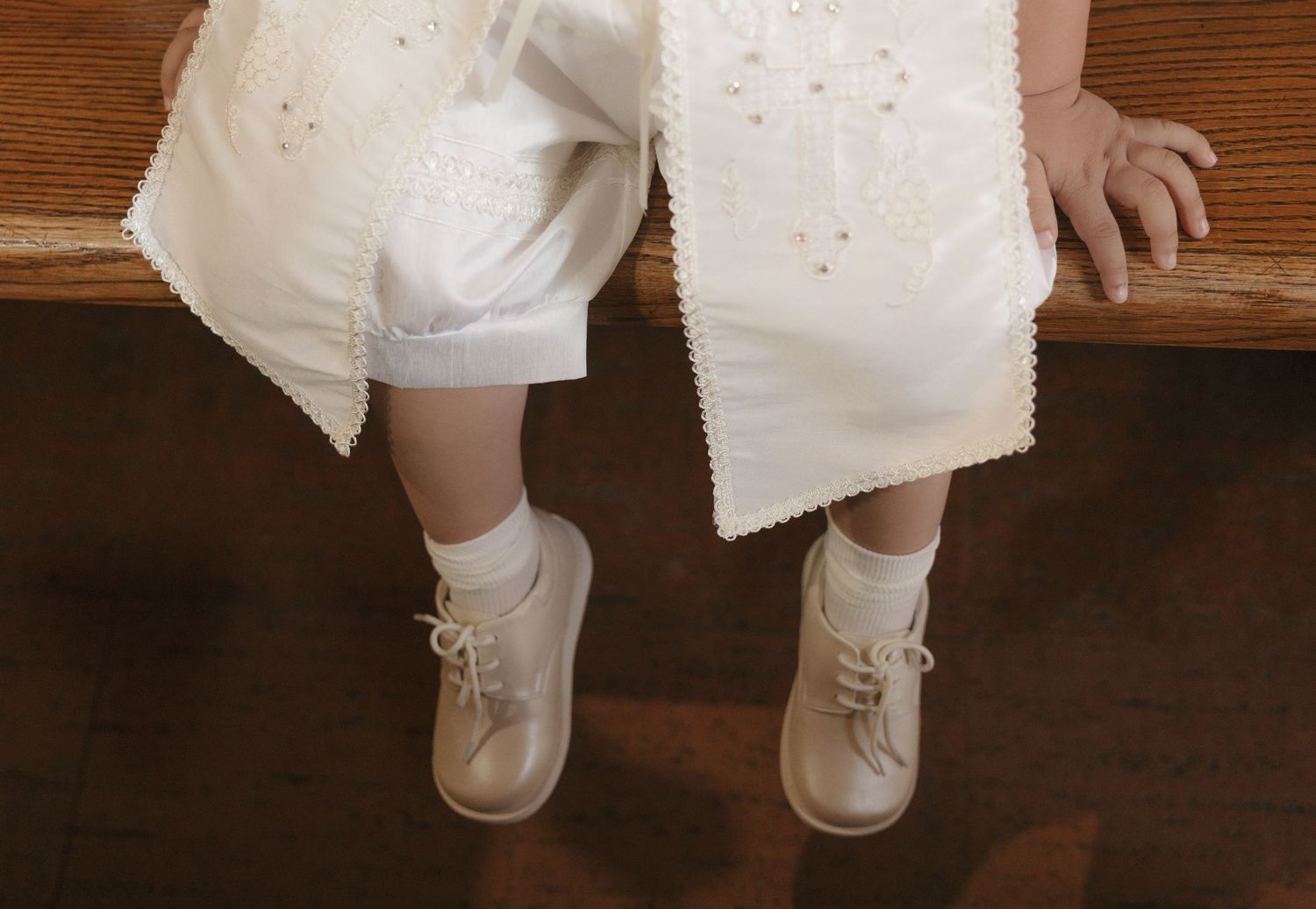Close-up of a toddler sitting on a bench wearing a white embroidered dress and cream lace-up ankle boots.