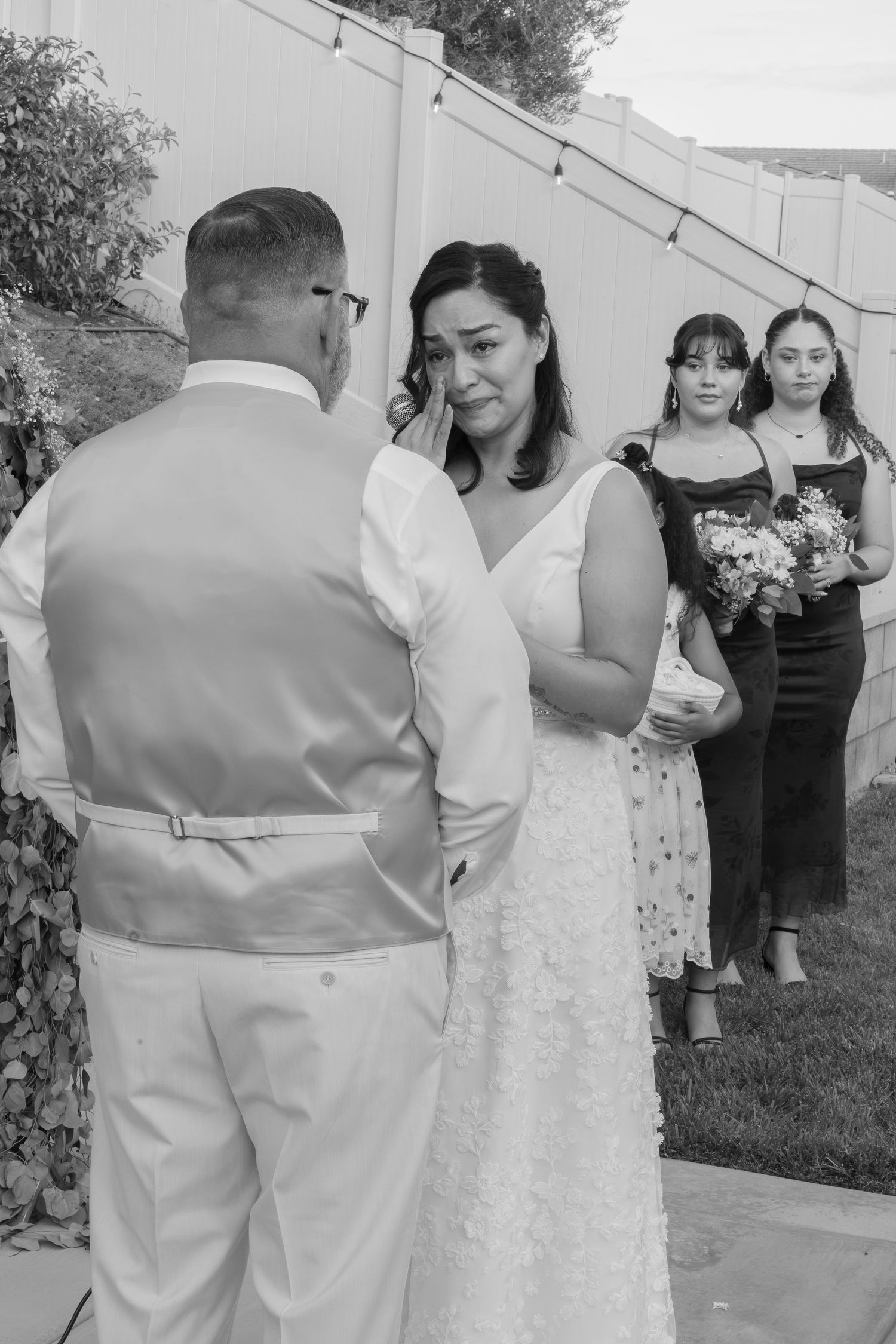 Emotional bride wipes tears during outdoor wedding ceremony while bridesmaids watch in background.