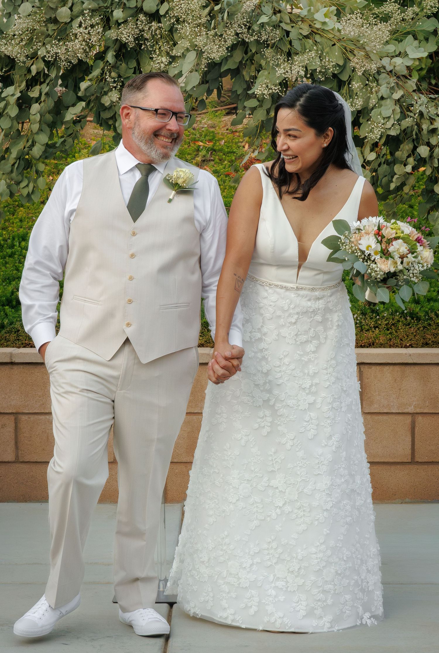 A smiling bride in a white floral gown holds a bouquet while a groom in a beige suit stands beside her outdoors.
