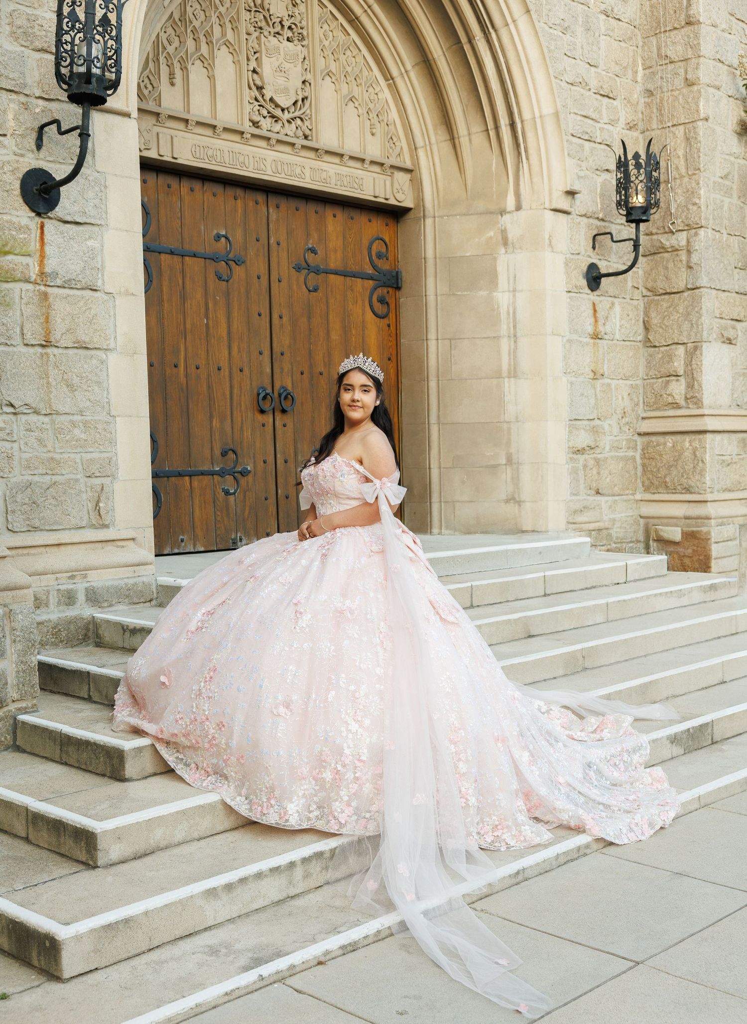 Latina quinceañera in blush pink ball gown with floral embroidery poses on gothic church steps.