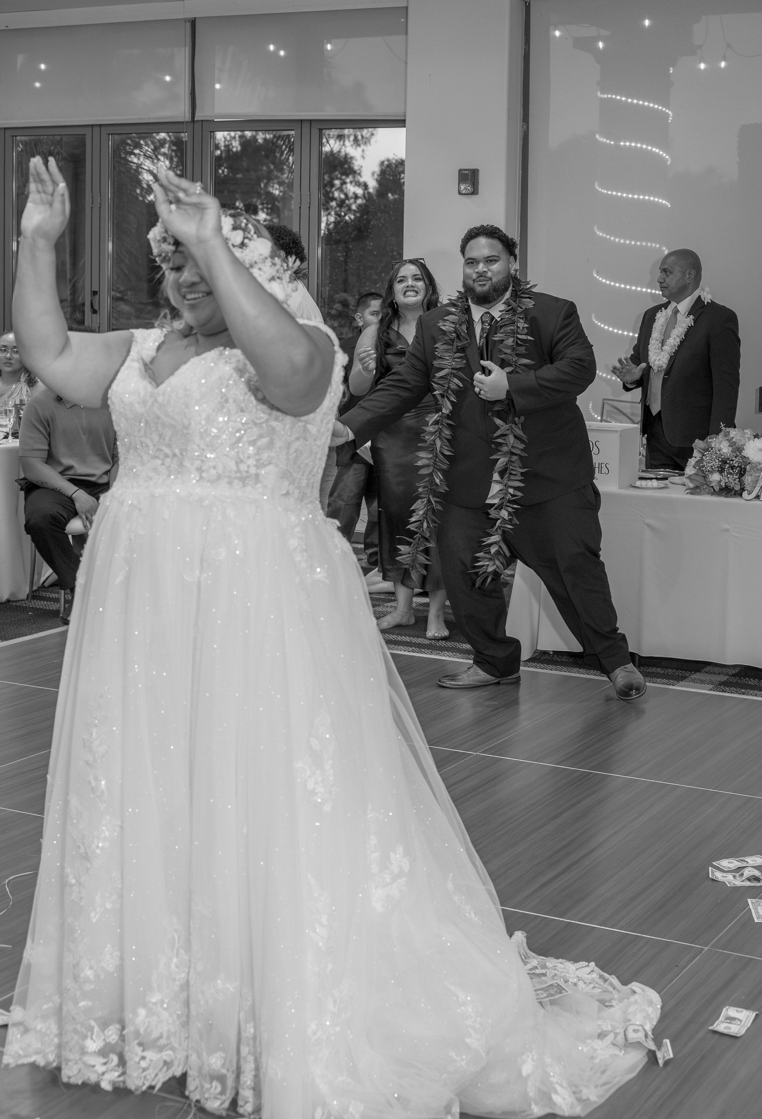 Bride in lace wedding gown raises arms on dance floor while guests celebrate at wedding reception.