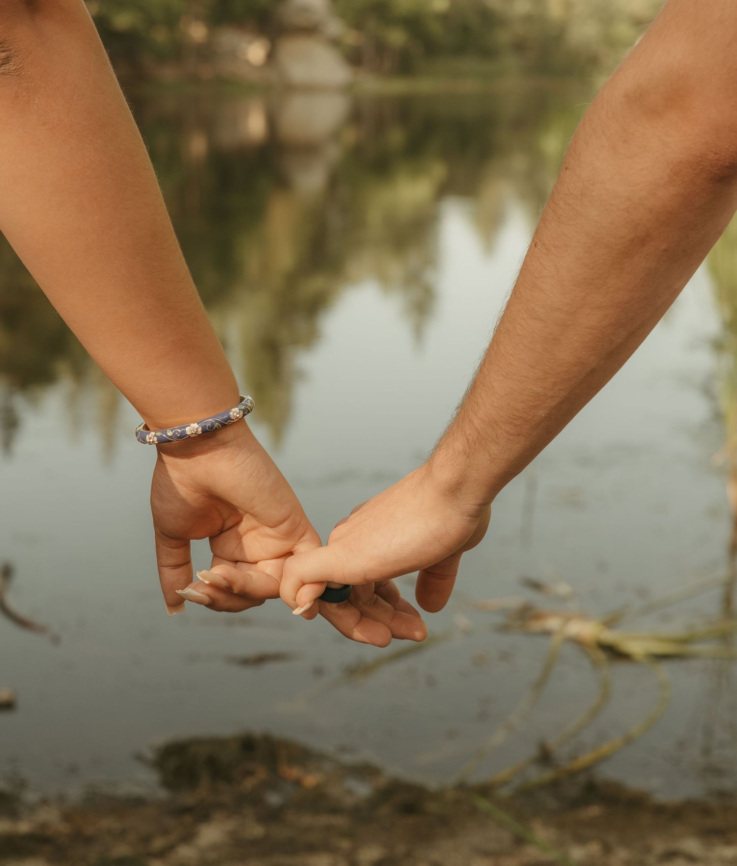 Couple holding hands near a reflective lake, one wrist adorned with a silver bracelet, romantic outdoor setting.