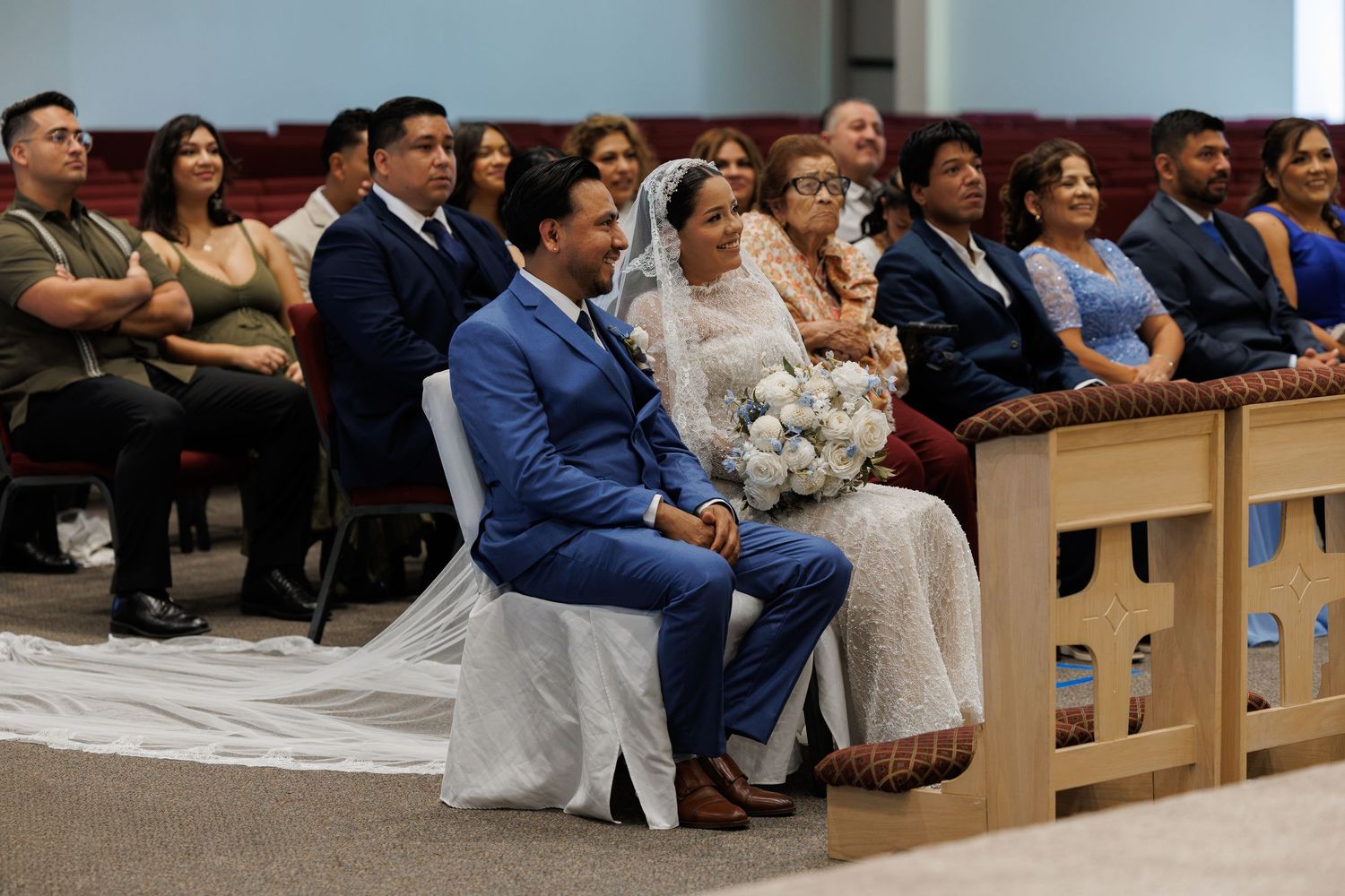 Bride in a white veil holding a bouquet sits beside her groom in a blue suit during a wedding ceremony as guests watch from the pews.