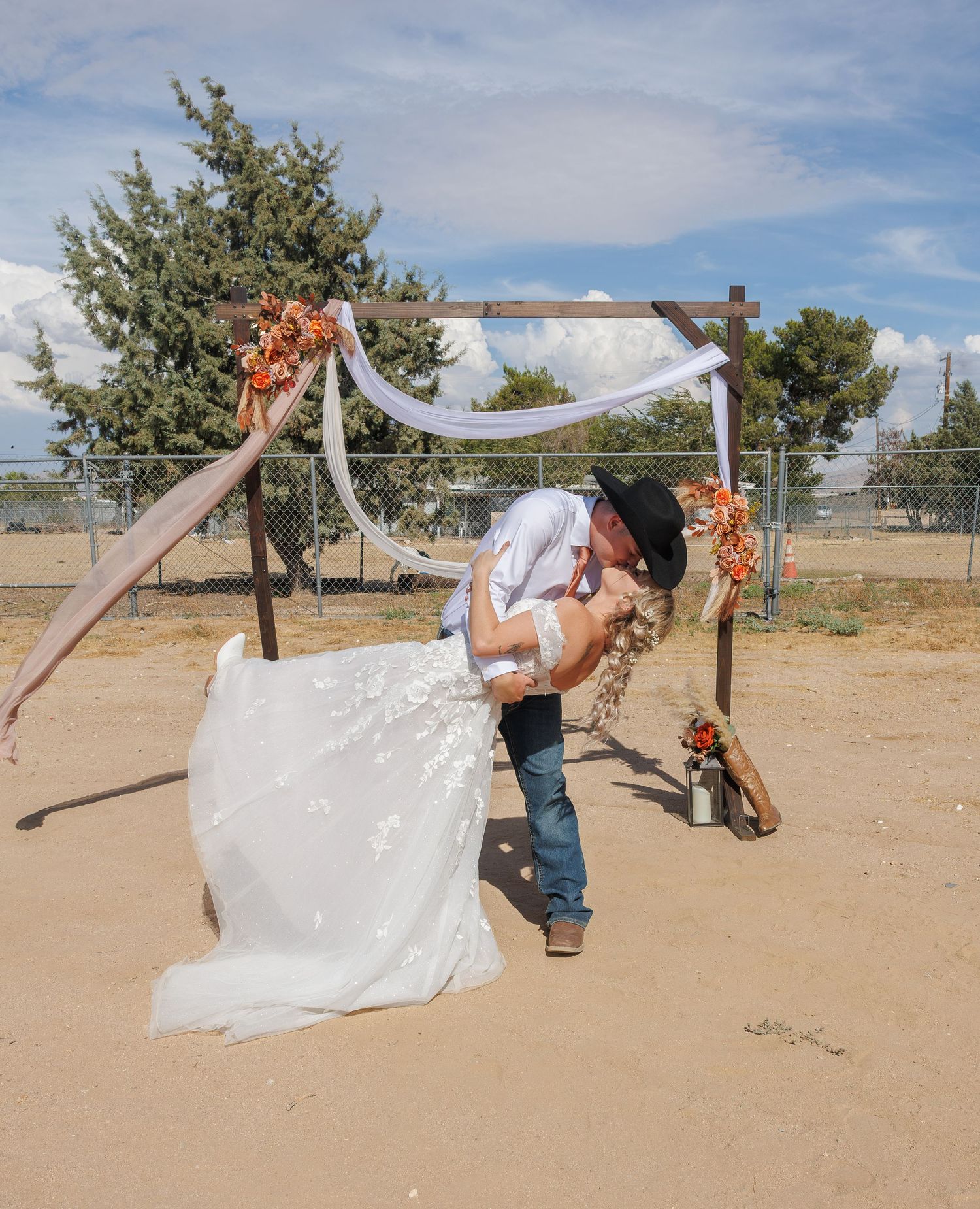 Bride and groom share romantic dip kiss under rustic wooden wedding arch outdoors.