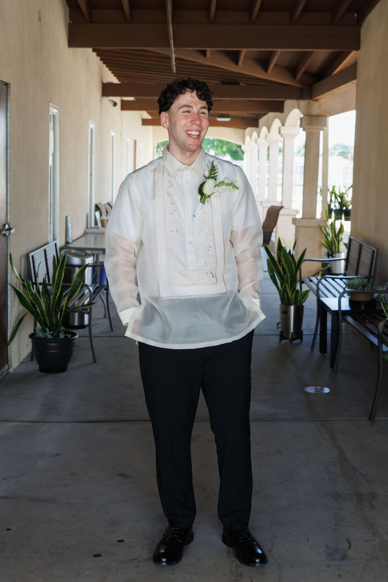Smiling groom in traditional barong tagalog with boutonniere stands at outdoor wedding venue.