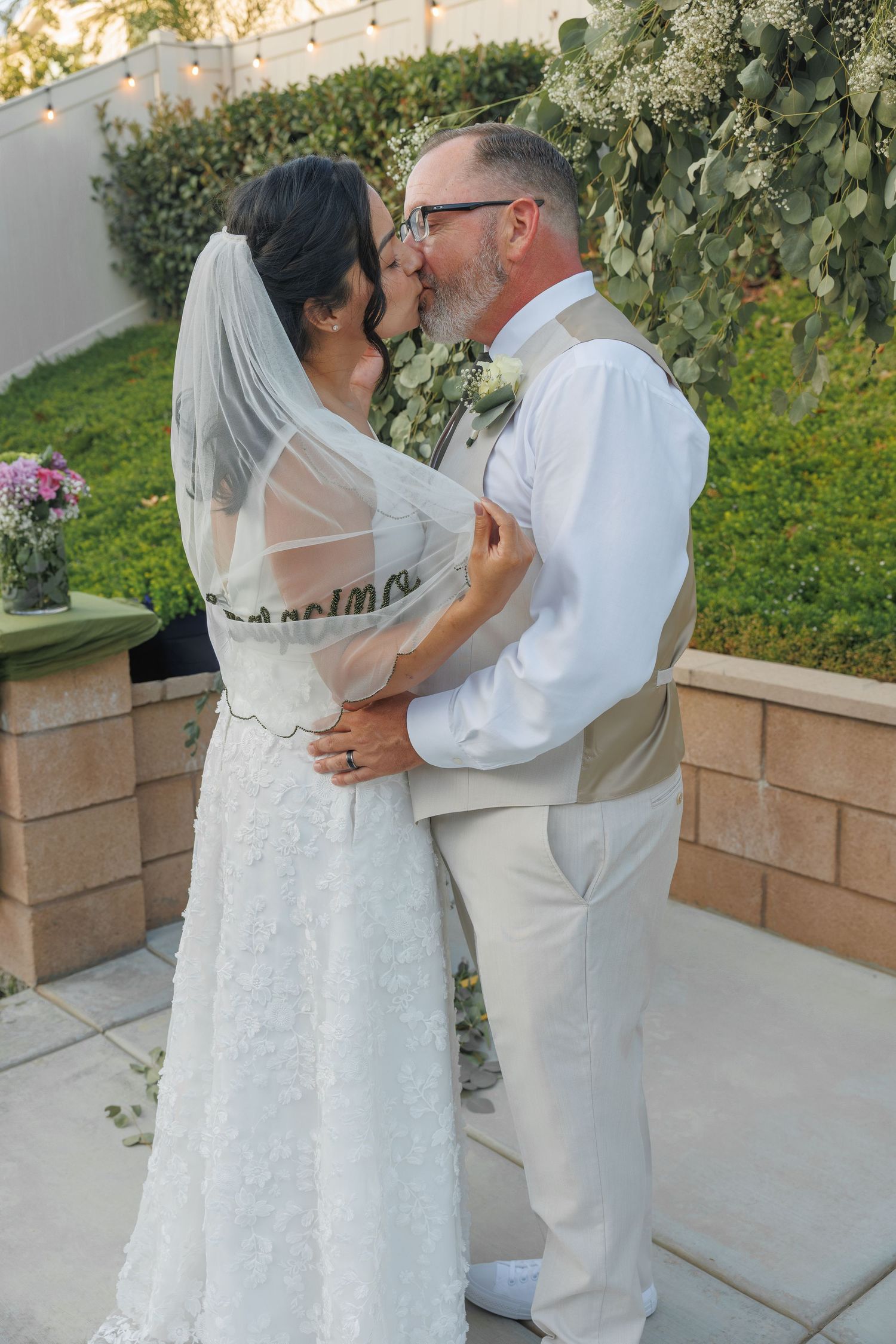 A bride and groom share their first kiss at an outdoor wedding ceremony, surrounded by lush greenery and string lights.