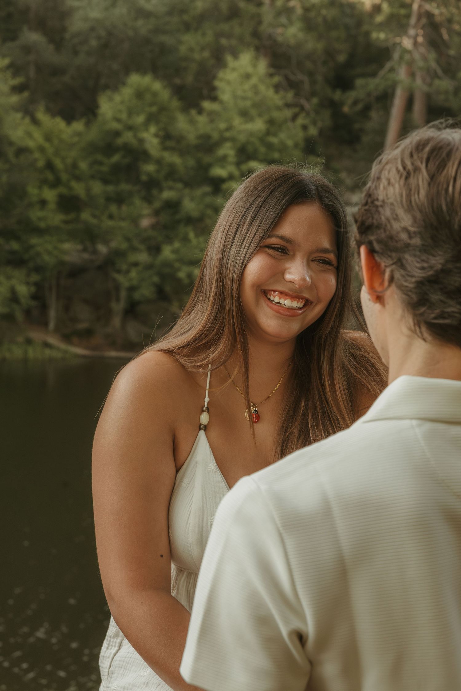 Happy young woman smiling at her partner outdoors near a lake with lush green trees behind her.