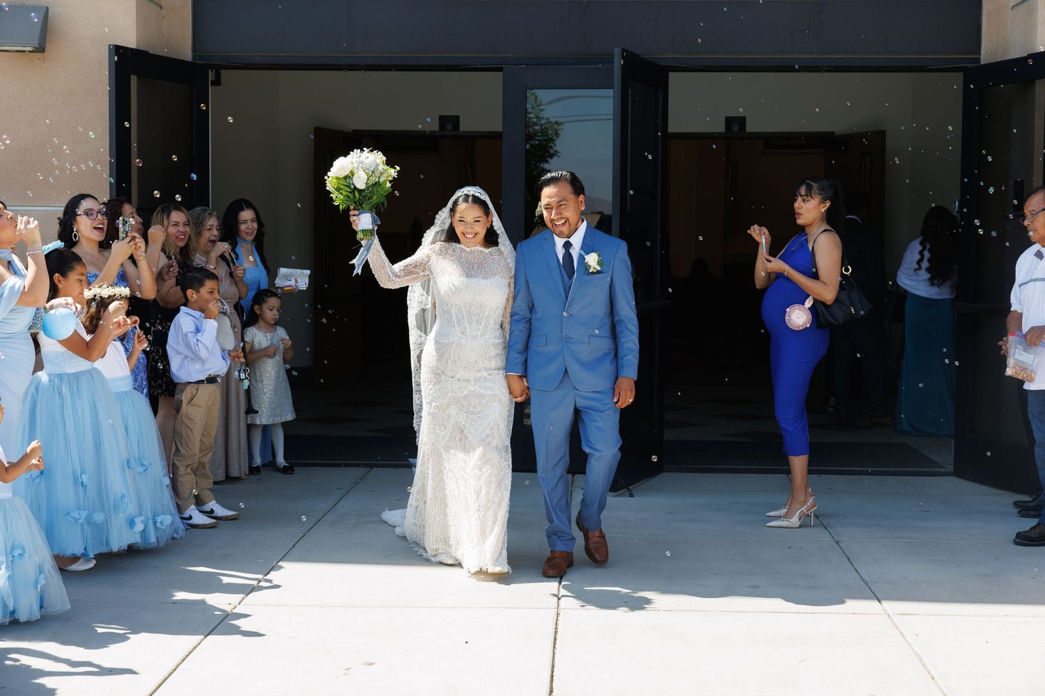 Bride in a white gown holding a bouquet walks hand in hand with a groom in a blue suit outside a church as guests in blue dresses cheer.
