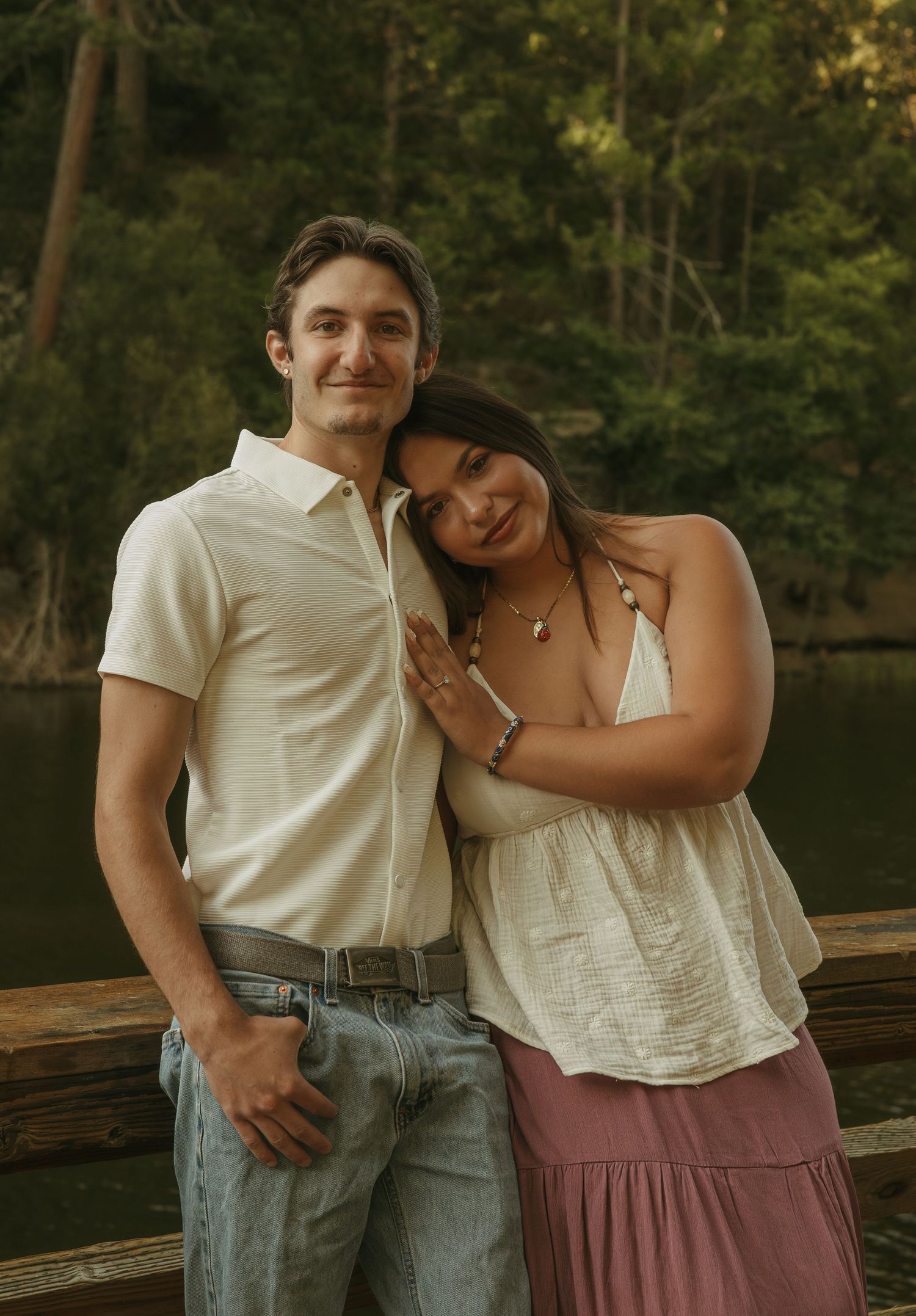 A couple poses on a wooden bridge near a lake, the woman leaning on the man, both wearing white tops.
