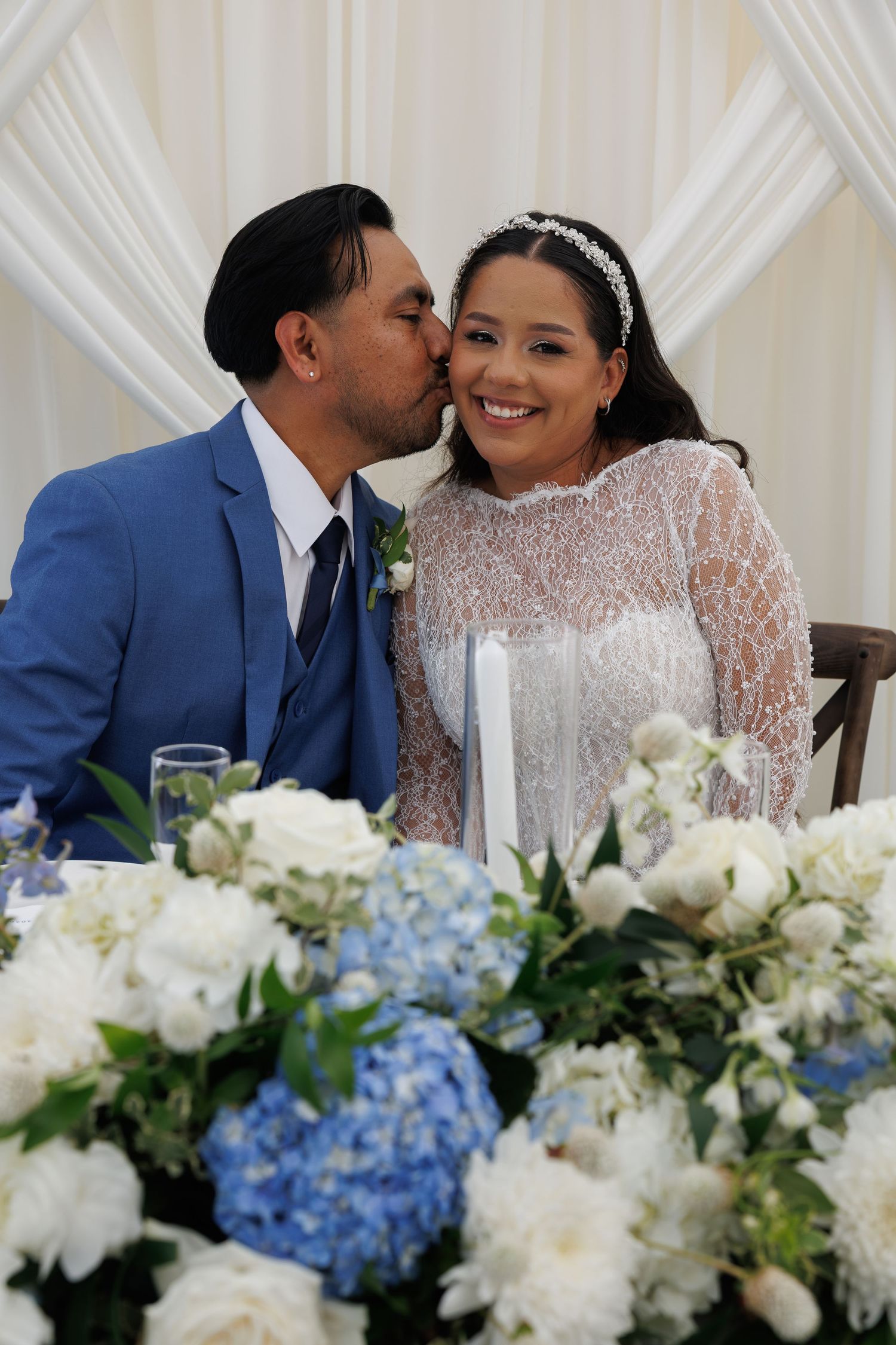 A groom in a blue suit kisses his smiling bride in a lace dress at the head table, surrounded by white and blue hydrangea centerpieces.