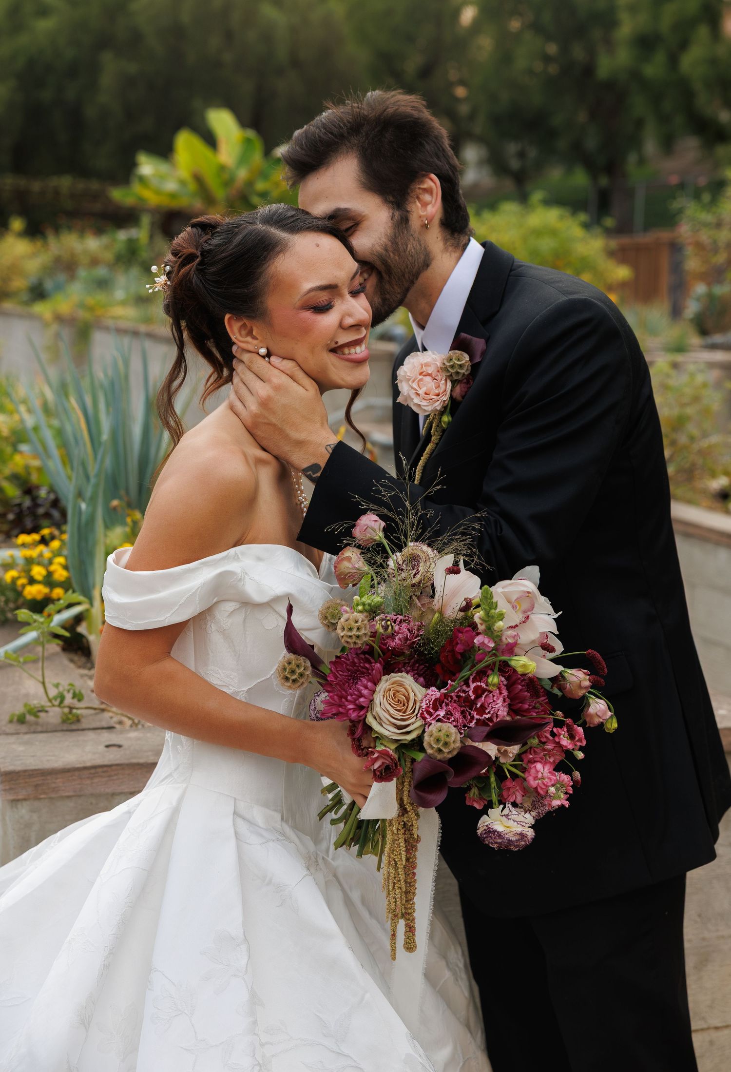 A bride and groom share a romantic moment in a garden, she holds a lush bouquet of pink and burgundy flowers.