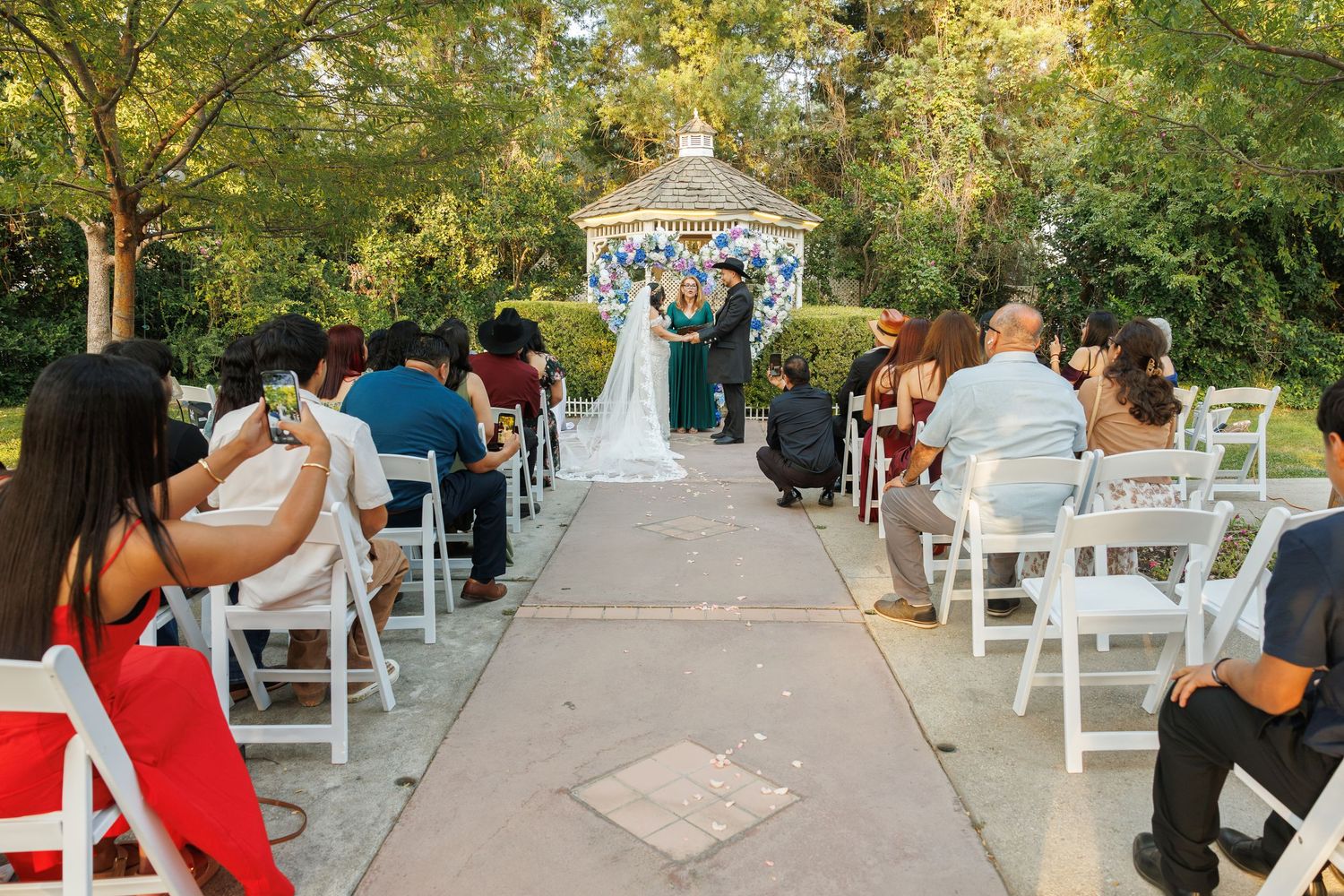 Guests seated in white chairs watch a couple exchange vows at a gazebo wedding ceremony outdoors.