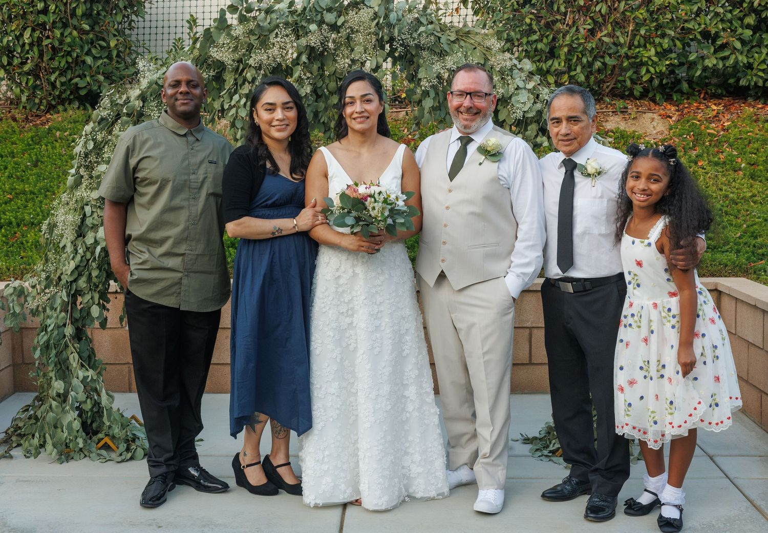 Wedding party group photo with bride and groom in white and tan suits under a lush greenery arch.