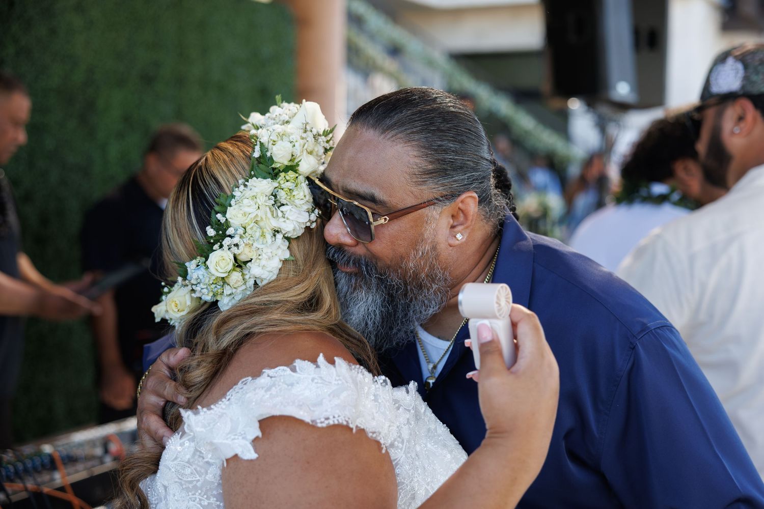 Bride and groom share a tender embrace at outdoor wedding reception, bride wearing floral crown with white roses.