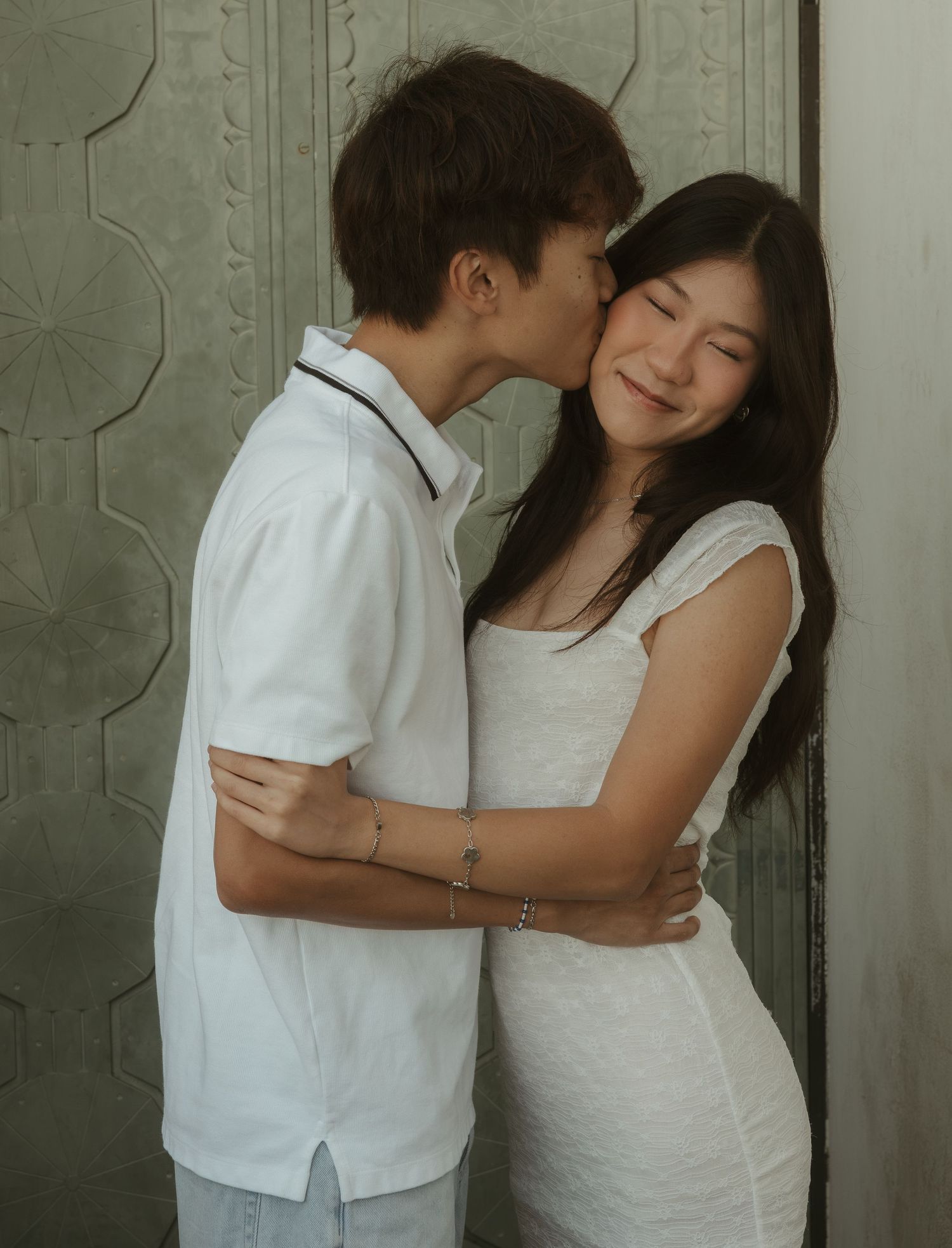 A young couple in white outfits share a tender moment, with the man kissing the woman's cheek as she smiles.