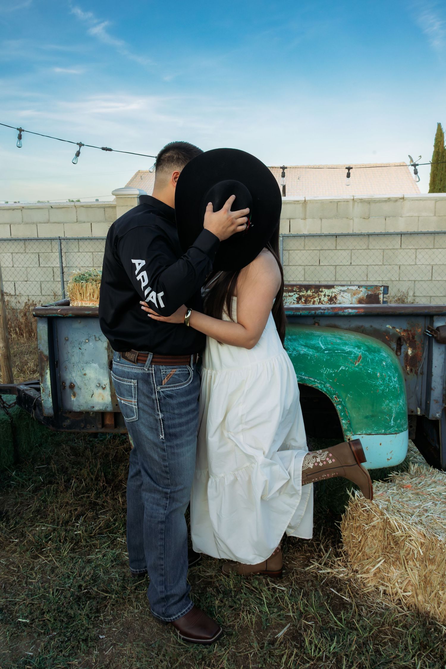Couple kissing behind a black cowboy hat, woman in white dress and boots, man in Ariat hoodie and jeans, near vintage truck.