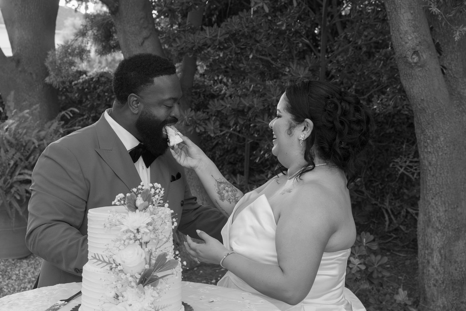 Bride feeding wedding cake to groom at outdoor reception table in black and white photo.