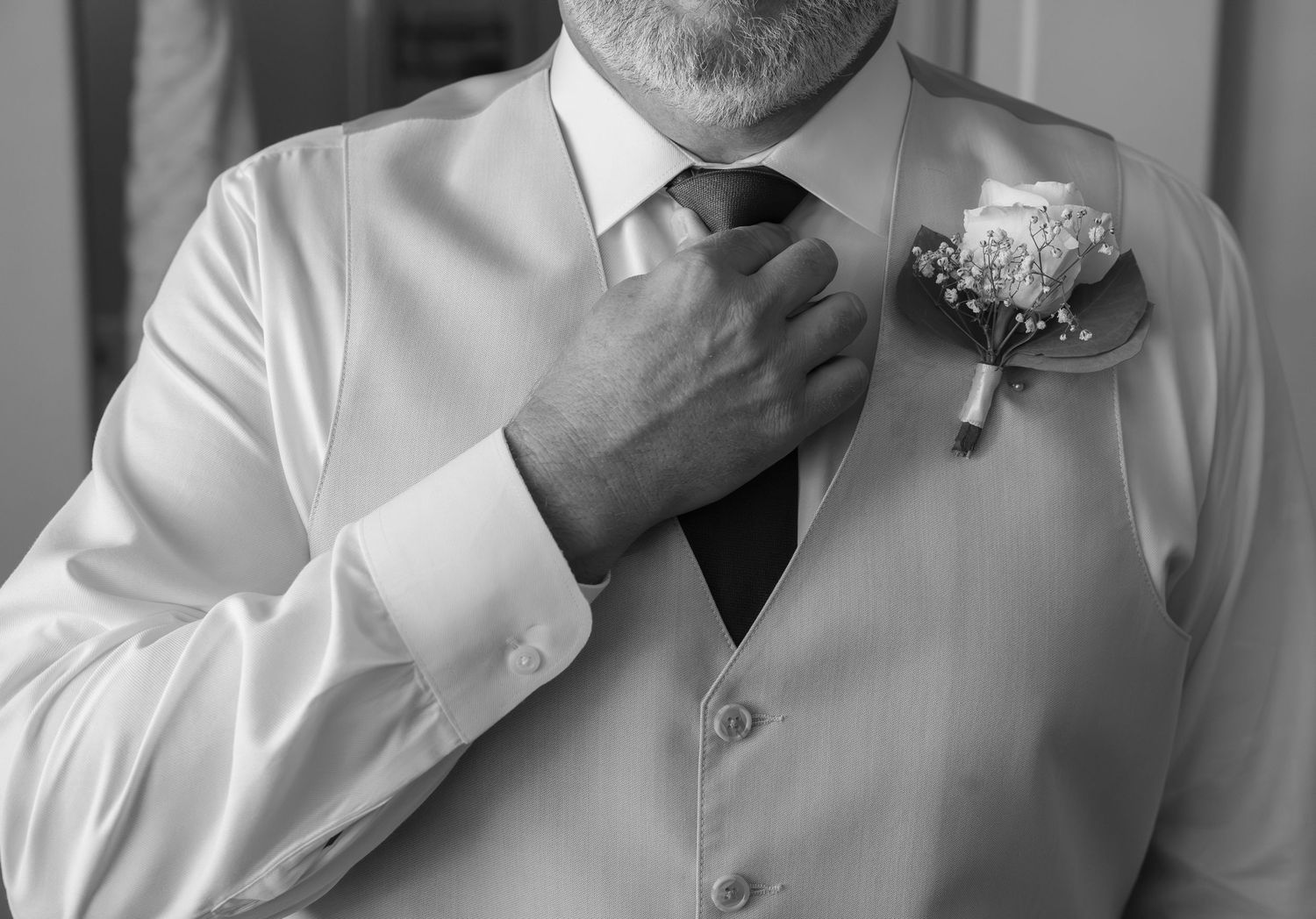 Black and white photo of a groom in a vest adjusting his tie with a boutonniere on his lapel.