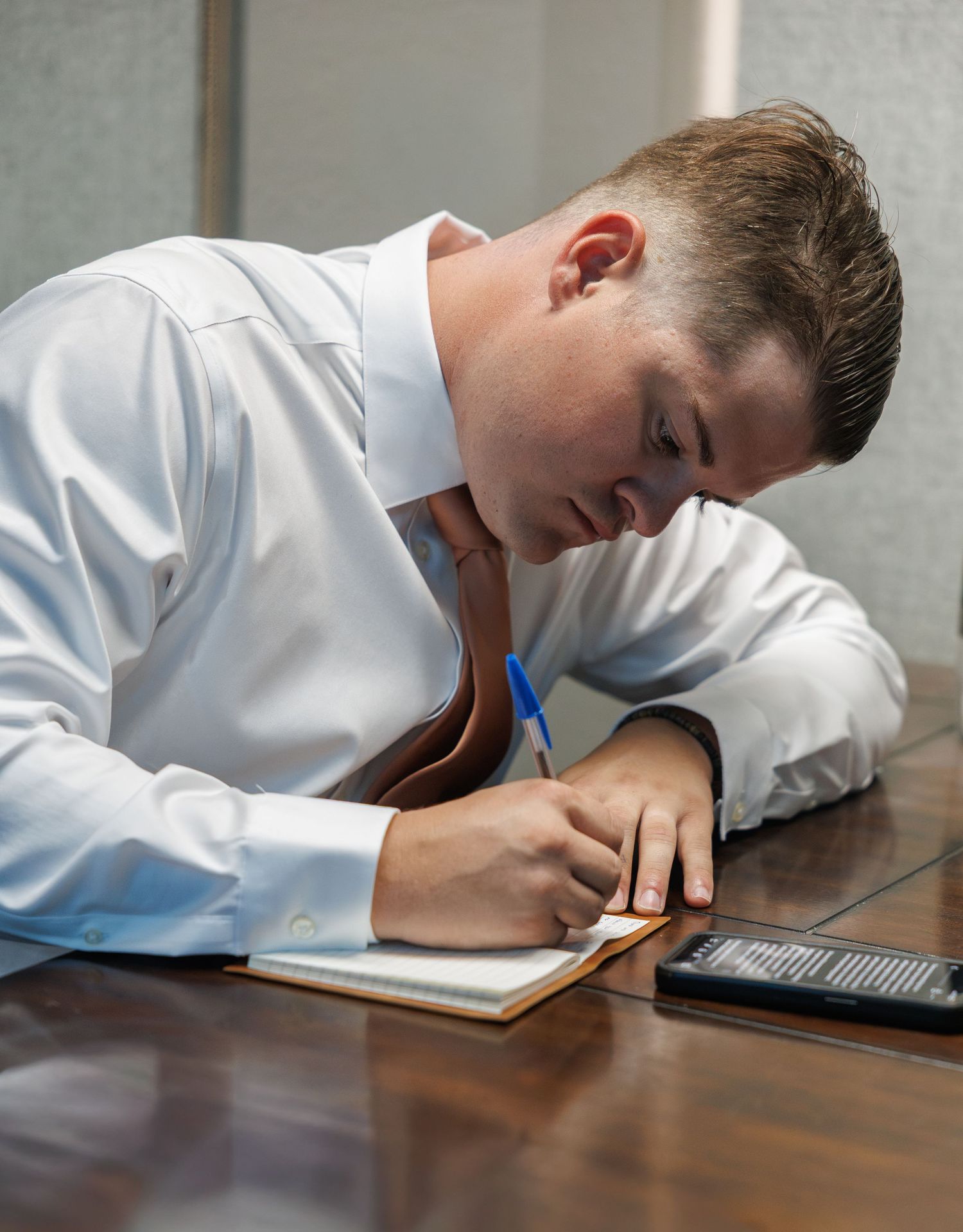 Young professional man in white dress shirt writing notes in a notebook at a wooden desk.