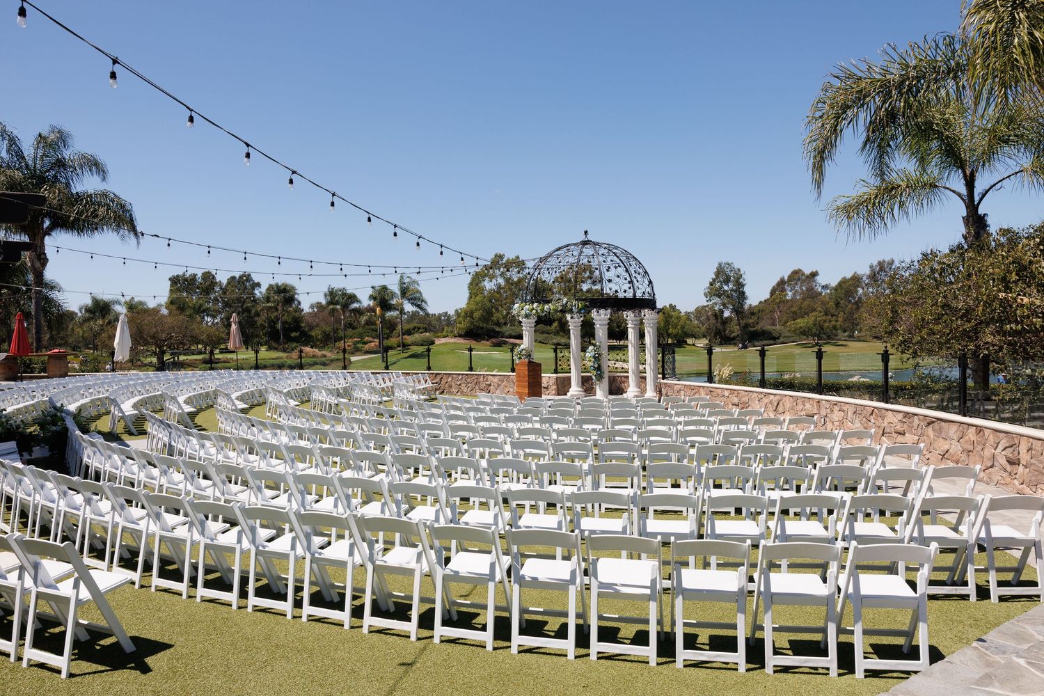 Outdoor wedding ceremony setup with white chairs arranged in rows facing an ornate gazebo on a sunny day.