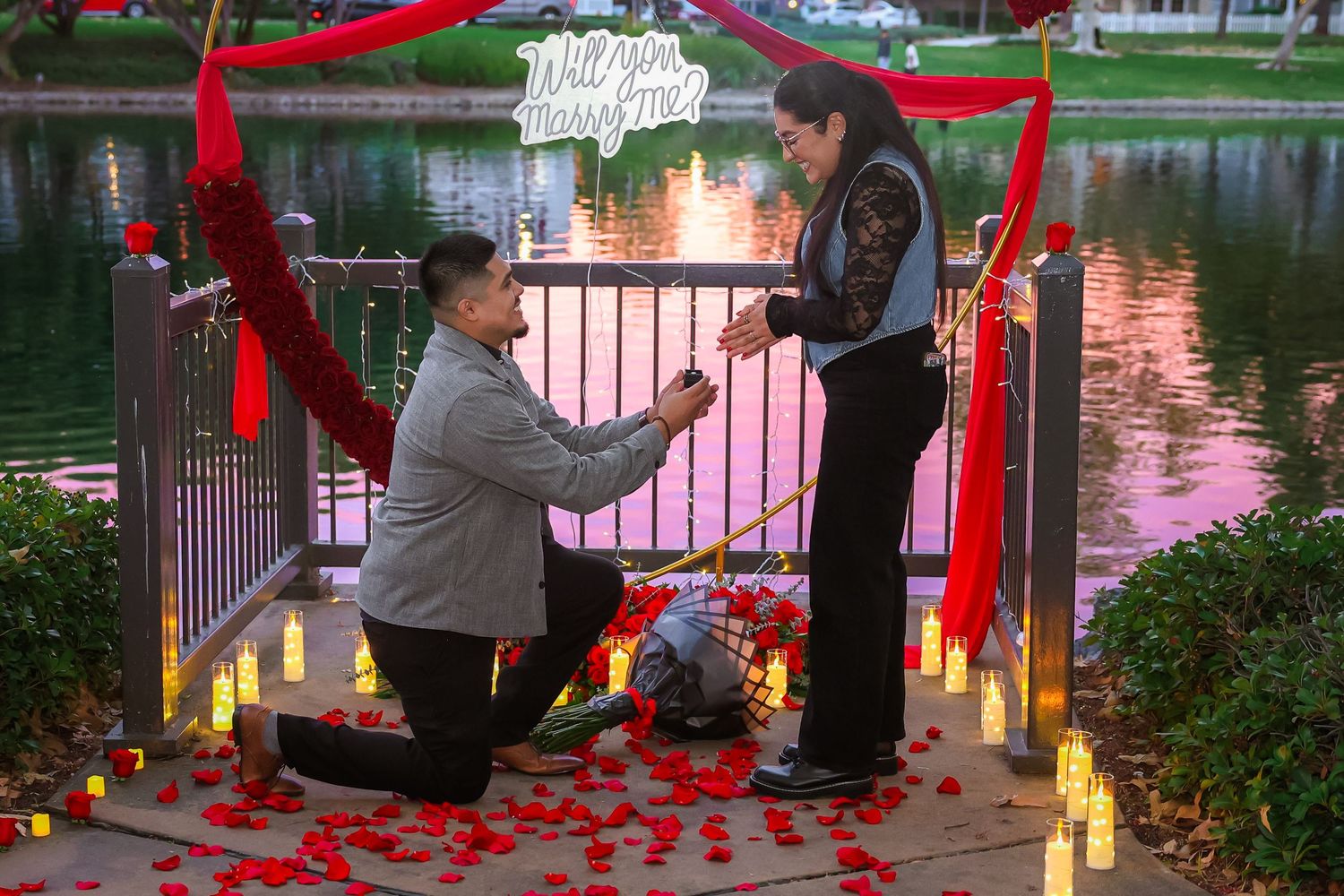 Romantic marriage proposal by a lake with rose petals, candles, and a Will You Marry Me sign at dusk.