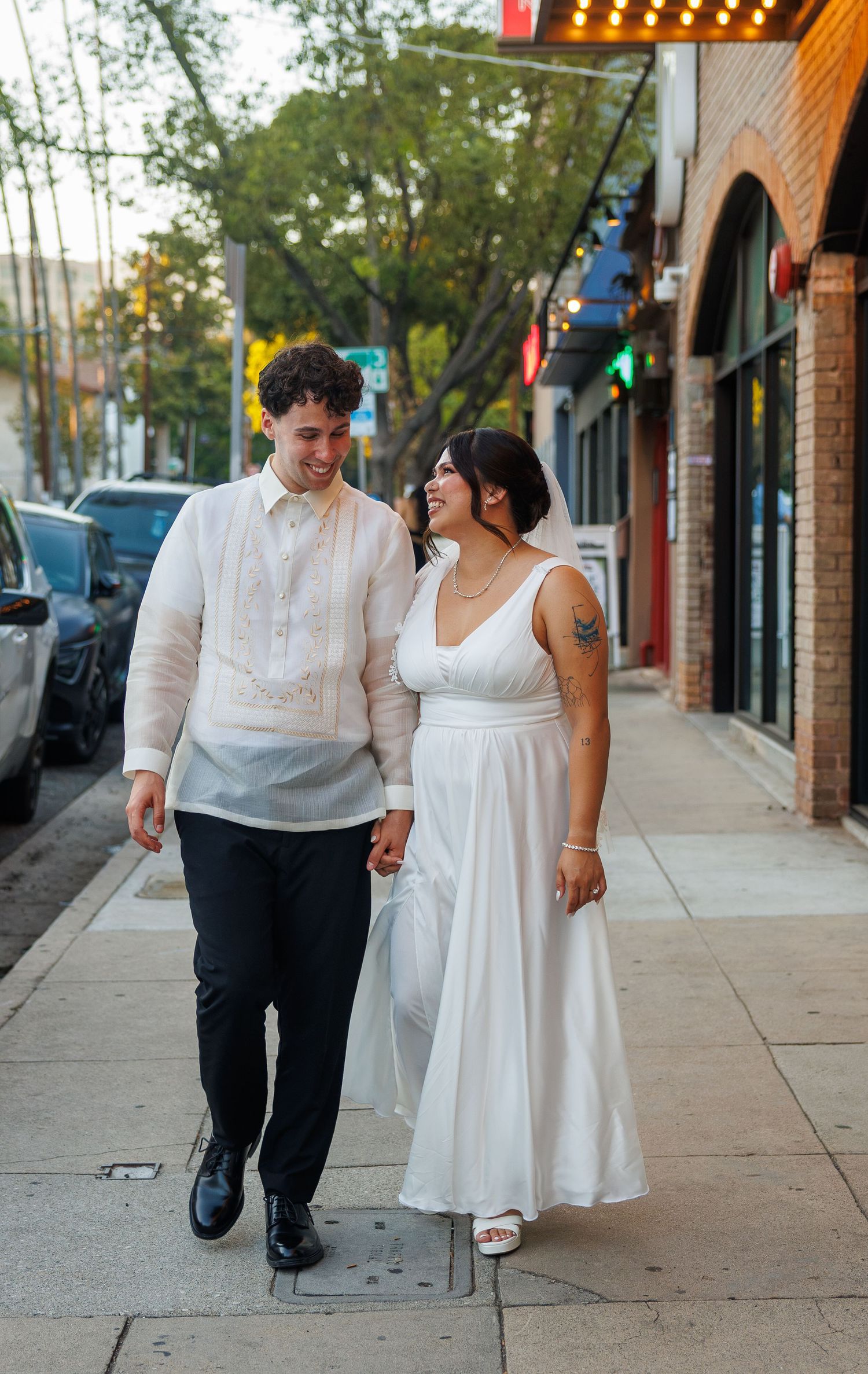 Newlywed couple walking hand-in-hand on a city sidewalk, groom in barong tagalog, bride in white midi dress.