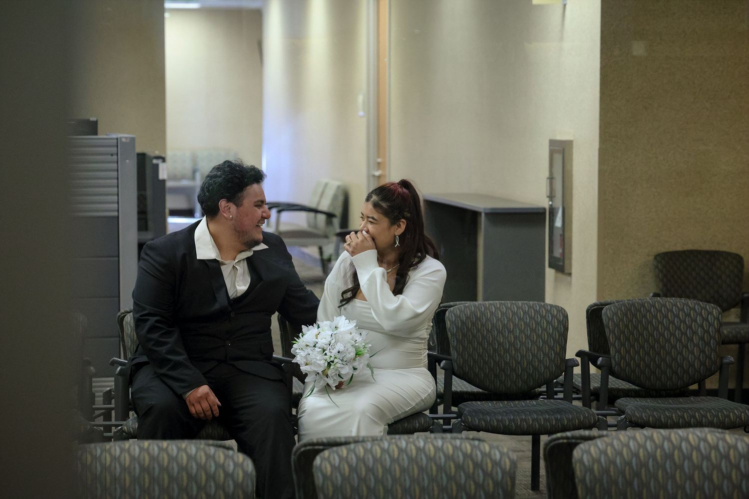 Bride laughing with groom while seated in courthouse waiting area holding white wedding bouquet.