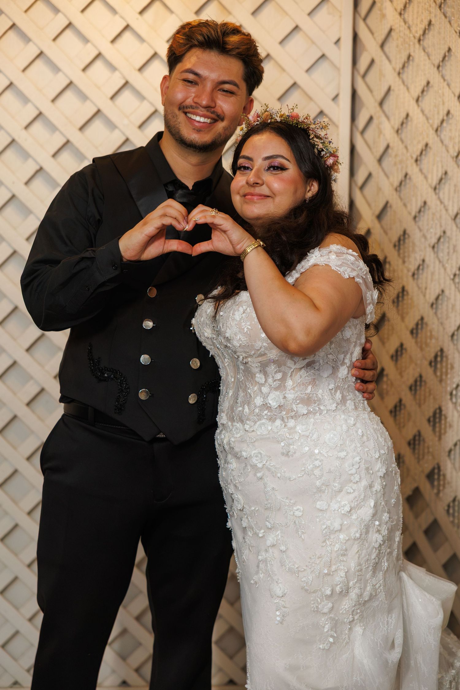 Smiling bride and groom make heart shape with hands, bride in lace gown with floral crown, groom in black suit.