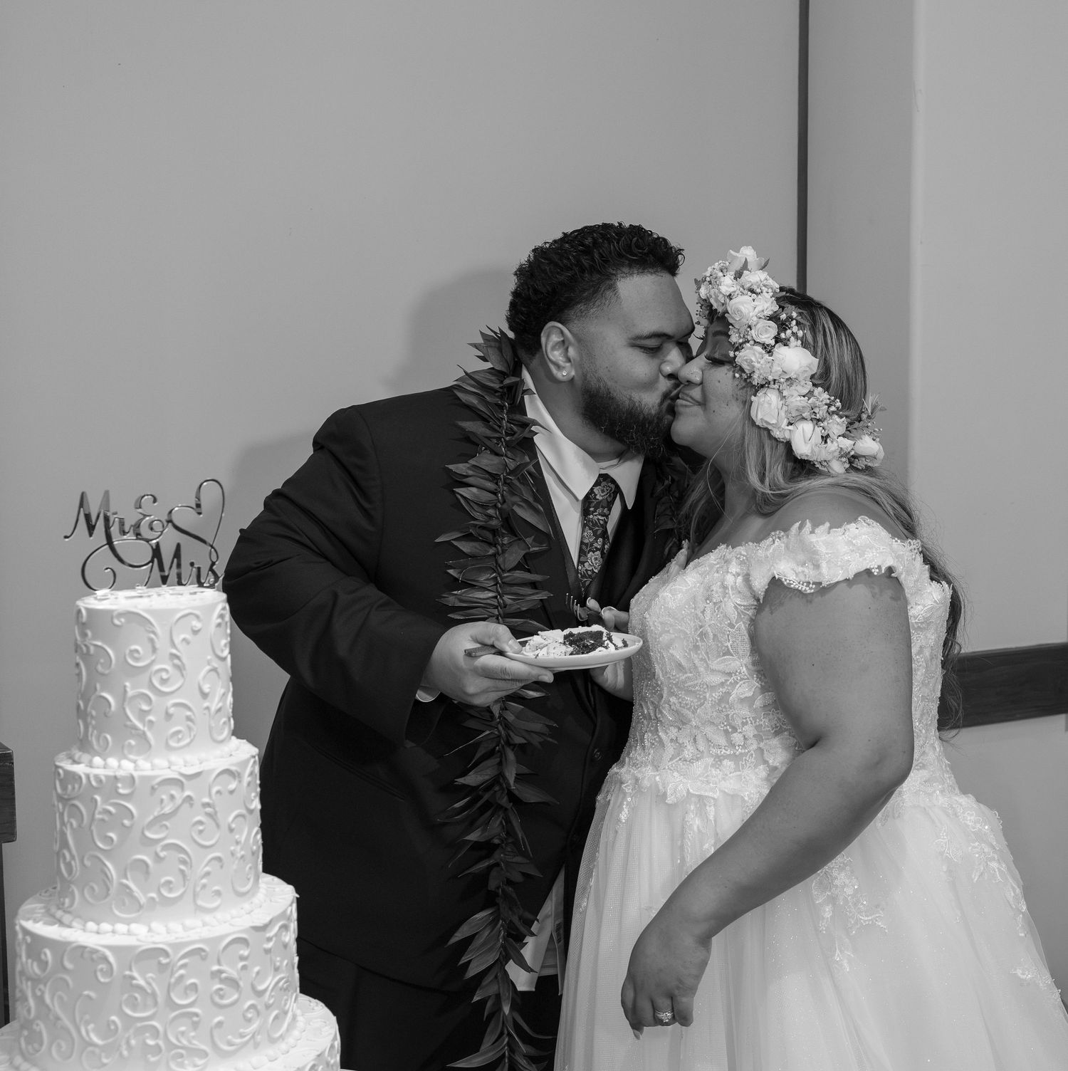 Black and white photo of newlyweds kissing beside a tiered wedding cake, bride in floral crown, groom in lei.