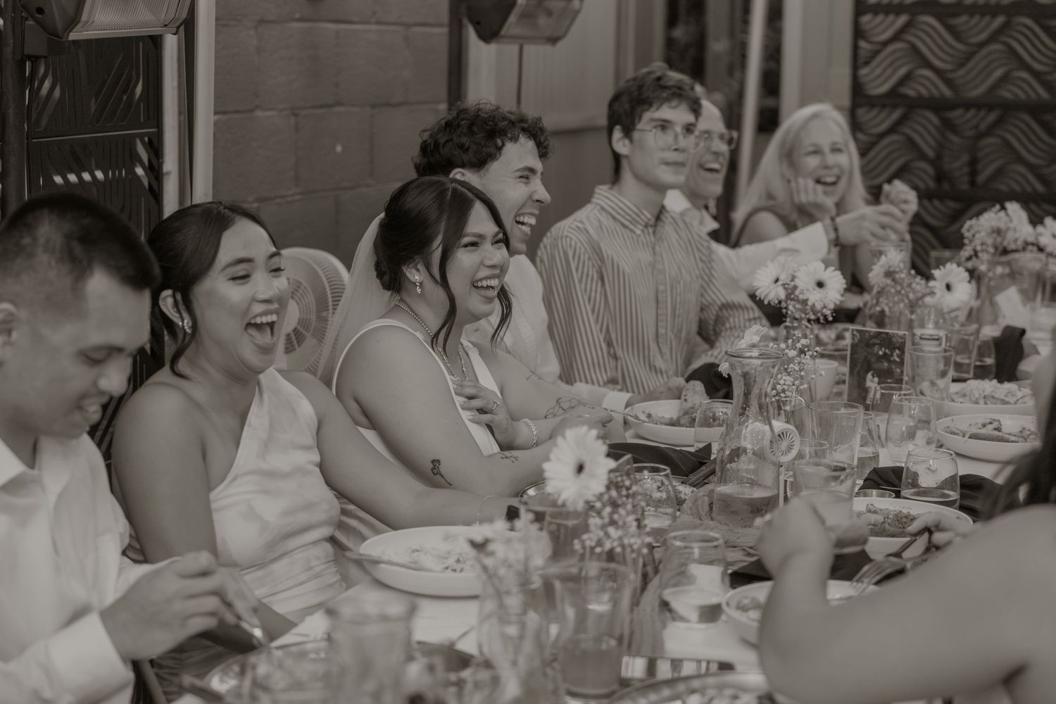 Black and white photo of wedding guests laughing at a reception dinner table with floral centerpieces.