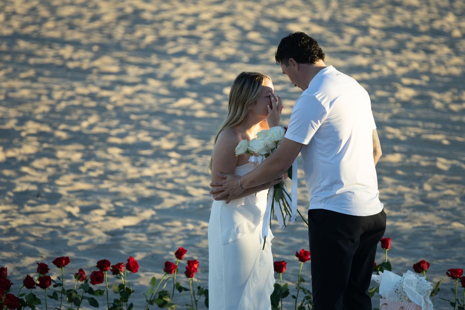 Couple sharing romantic moment on beach surrounded by red roses at sunset, bride in white dress holding bouquet.