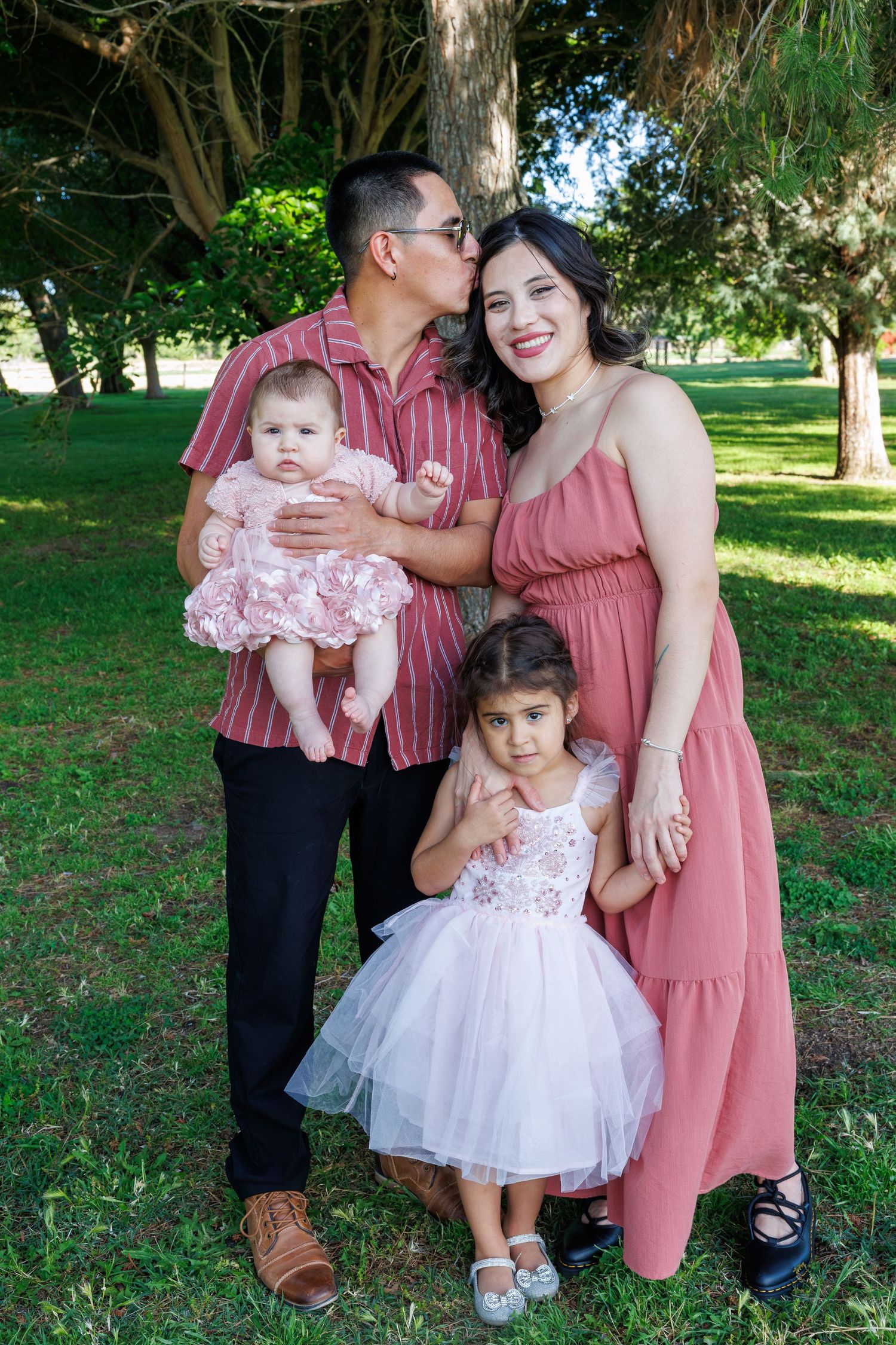 Family of four poses outdoors in a park, parents in pink outfits with two young daughters in white and pink dresses.