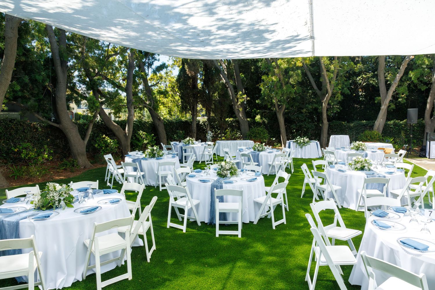 Elegant outdoor wedding reception with white round tables, folding chairs, and lush green lawn under a shaded canopy.
