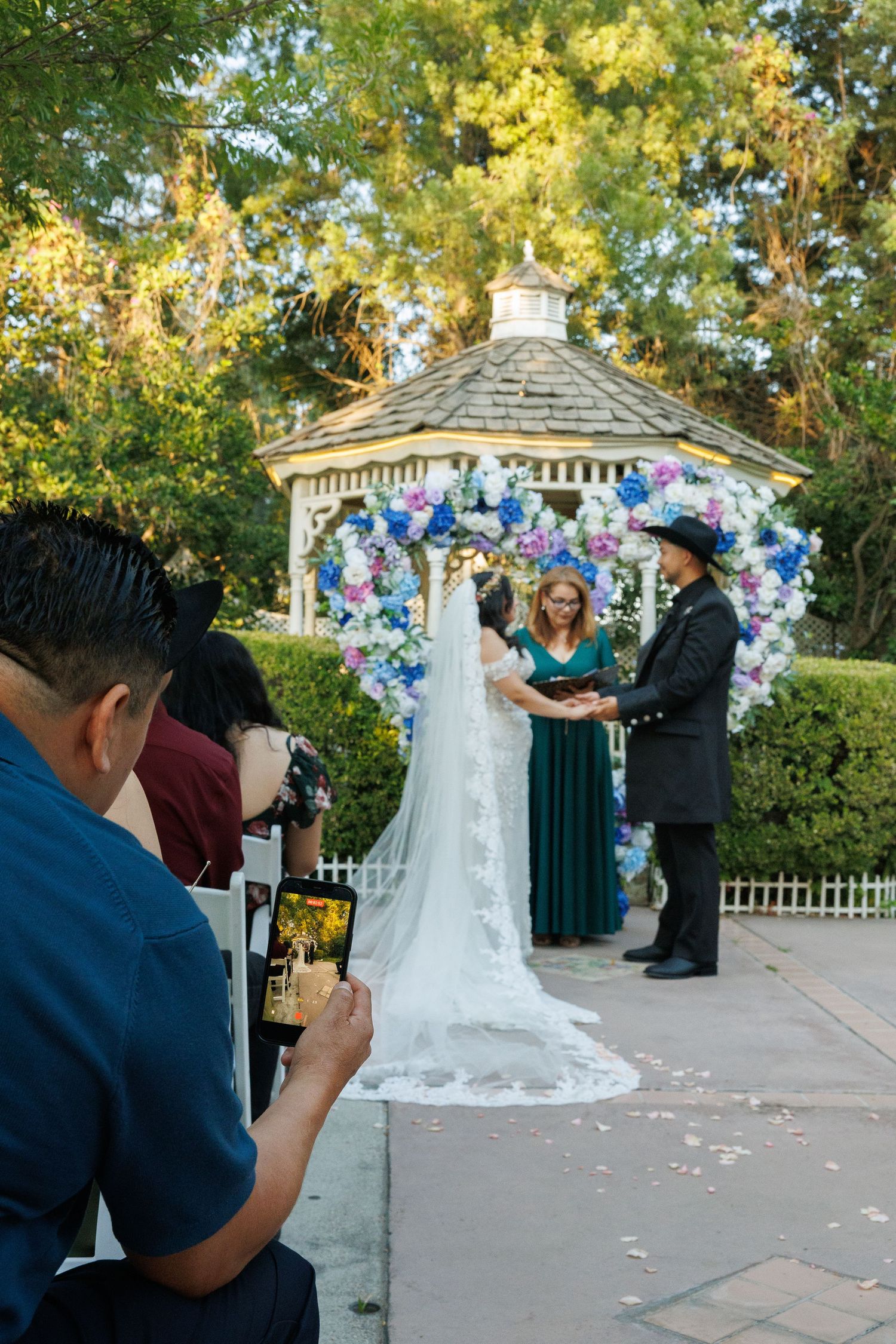 Outdoor wedding ceremony at a floral gazebo with bride, groom, and officiant while guests photograph the moment.