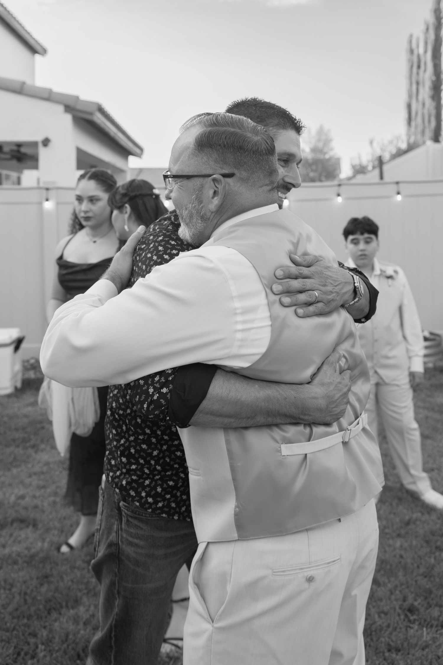 Black and white photo of two men sharing a heartfelt hug at an outdoor backyard celebration.