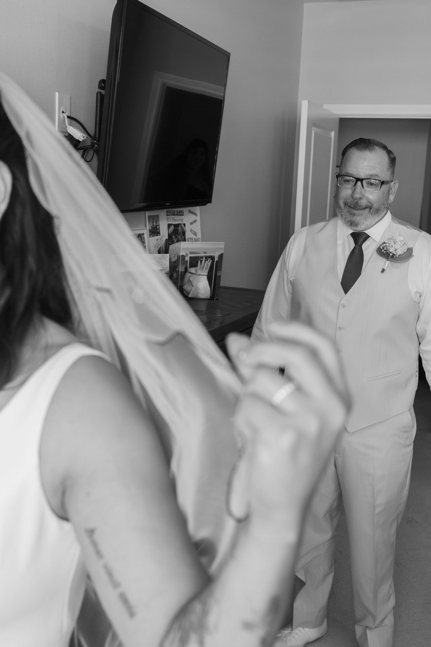 Black and white photo of a bride holding her veil as a man in a suit watches during wedding preparations.