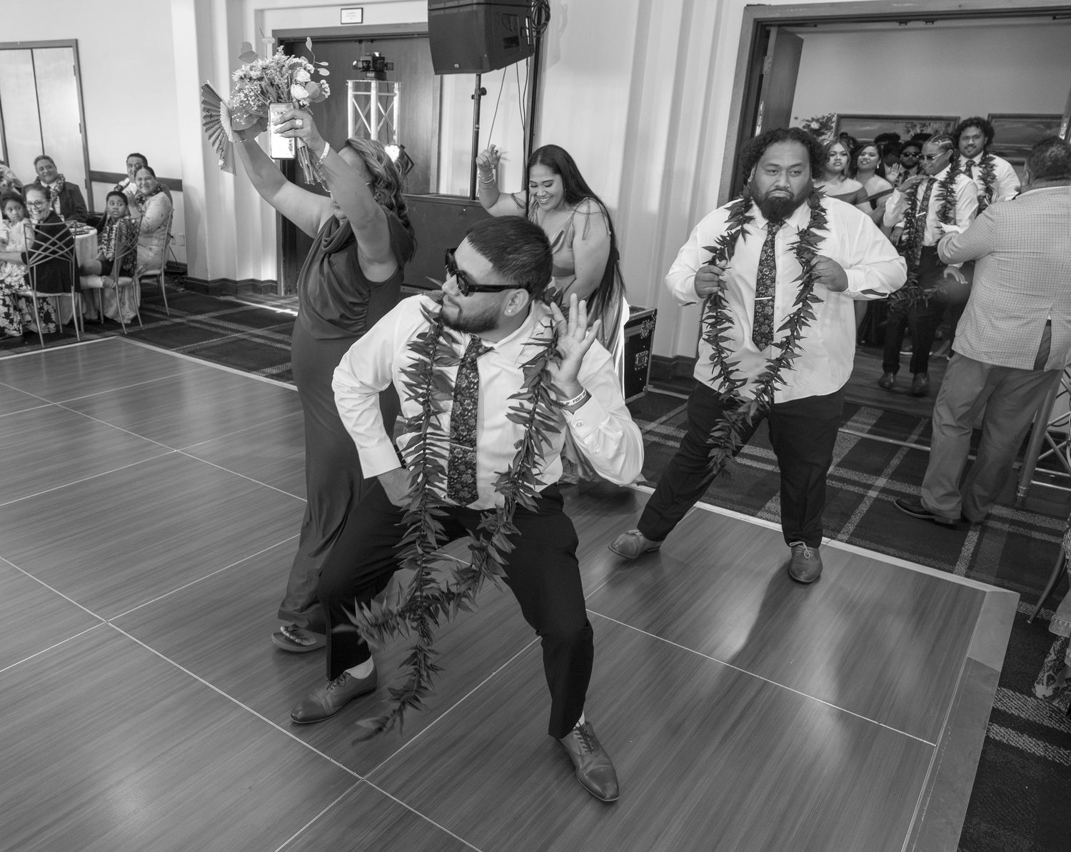 Men wearing floral leis perform a traditional dance at a wedding reception in black and white photo.