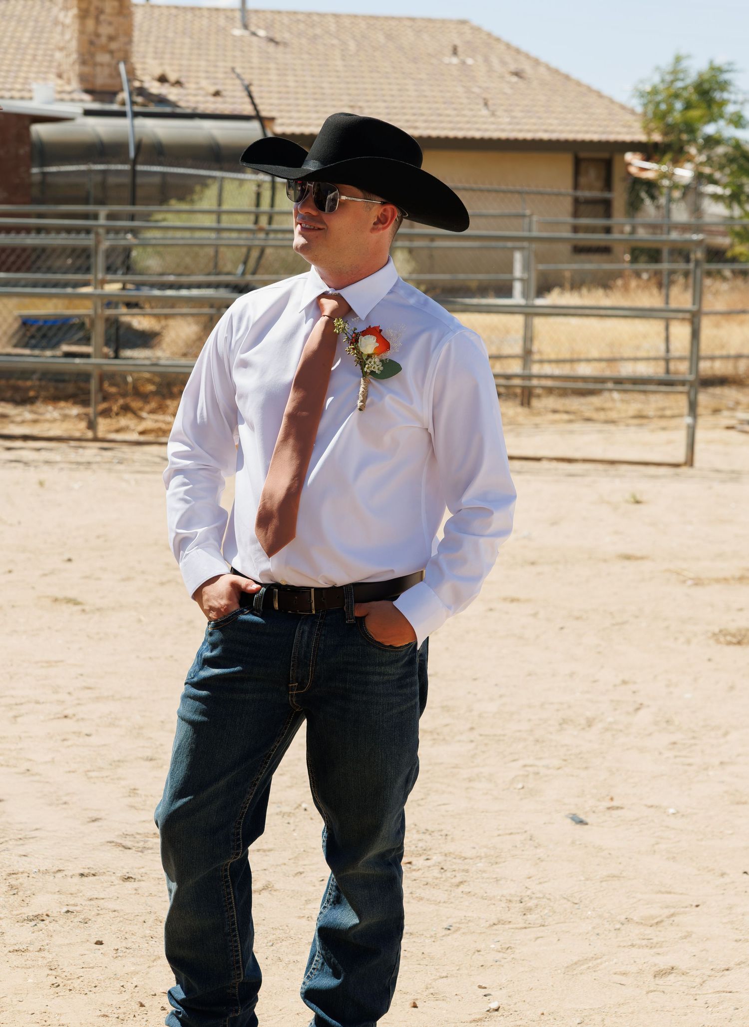Young man in cowboy hat, white shirt, brown tie, and jeans stands outdoors at a ranch event.