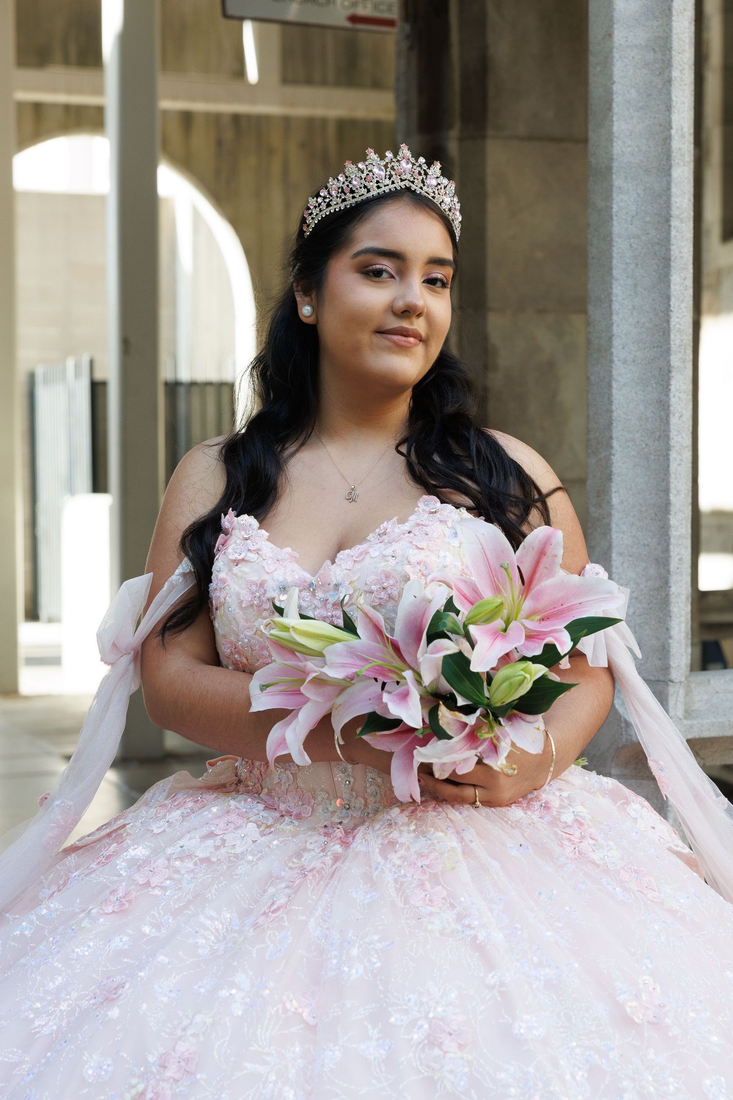 Young Latina woman in pink floral quinceañera ball gown with tiara holds pink lily bouquet.