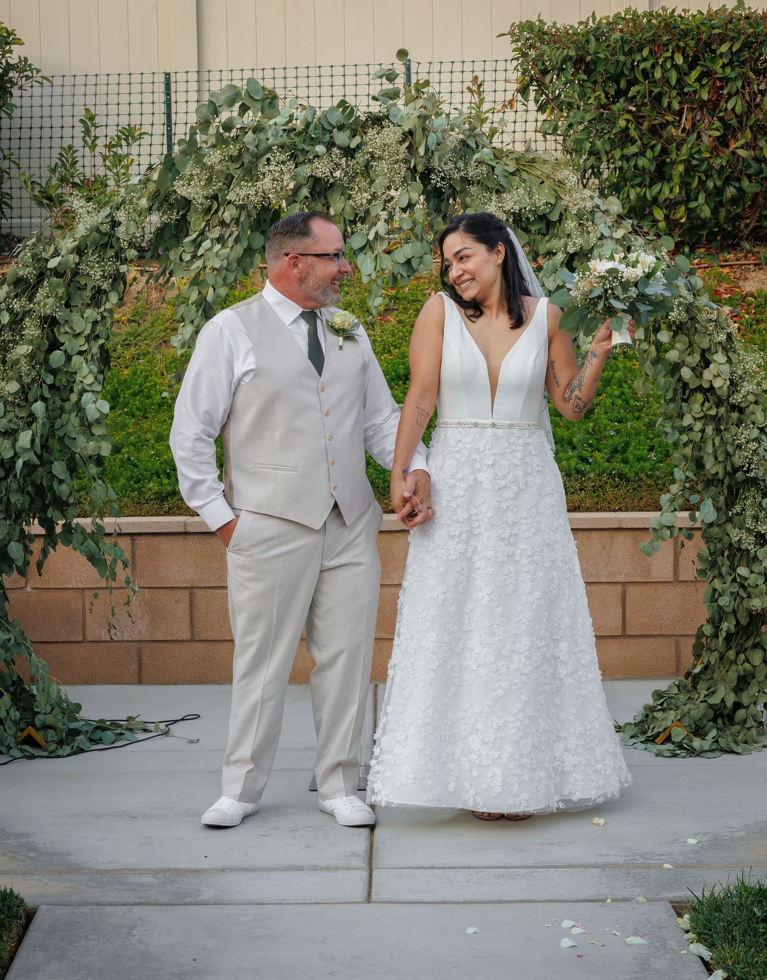 Bride and groom holding hands under a lush greenery arch, smiling at each other on their wedding day.