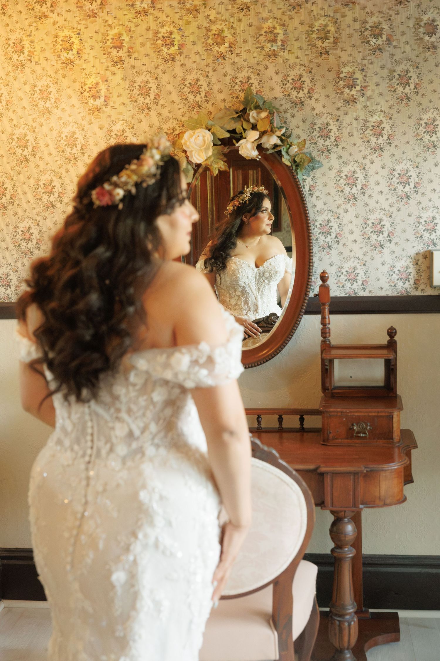 Bride in lace off-shoulder gown with floral crown admires her reflection in an ornate vintage mirror.