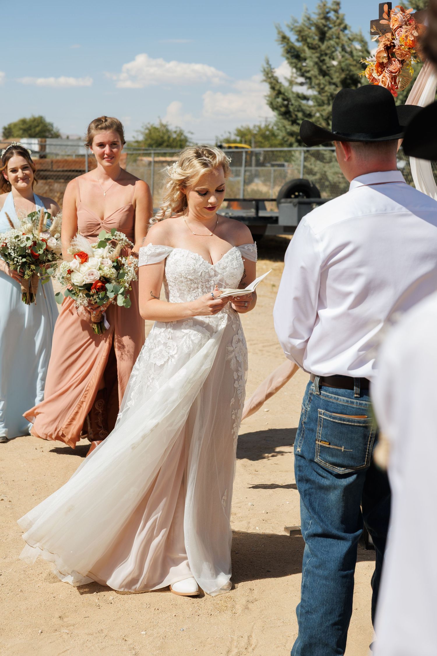 Radiant bride in off-shoulder lace gown reading vows with bridesmaids holding bouquets at sunny outdoor wedding.