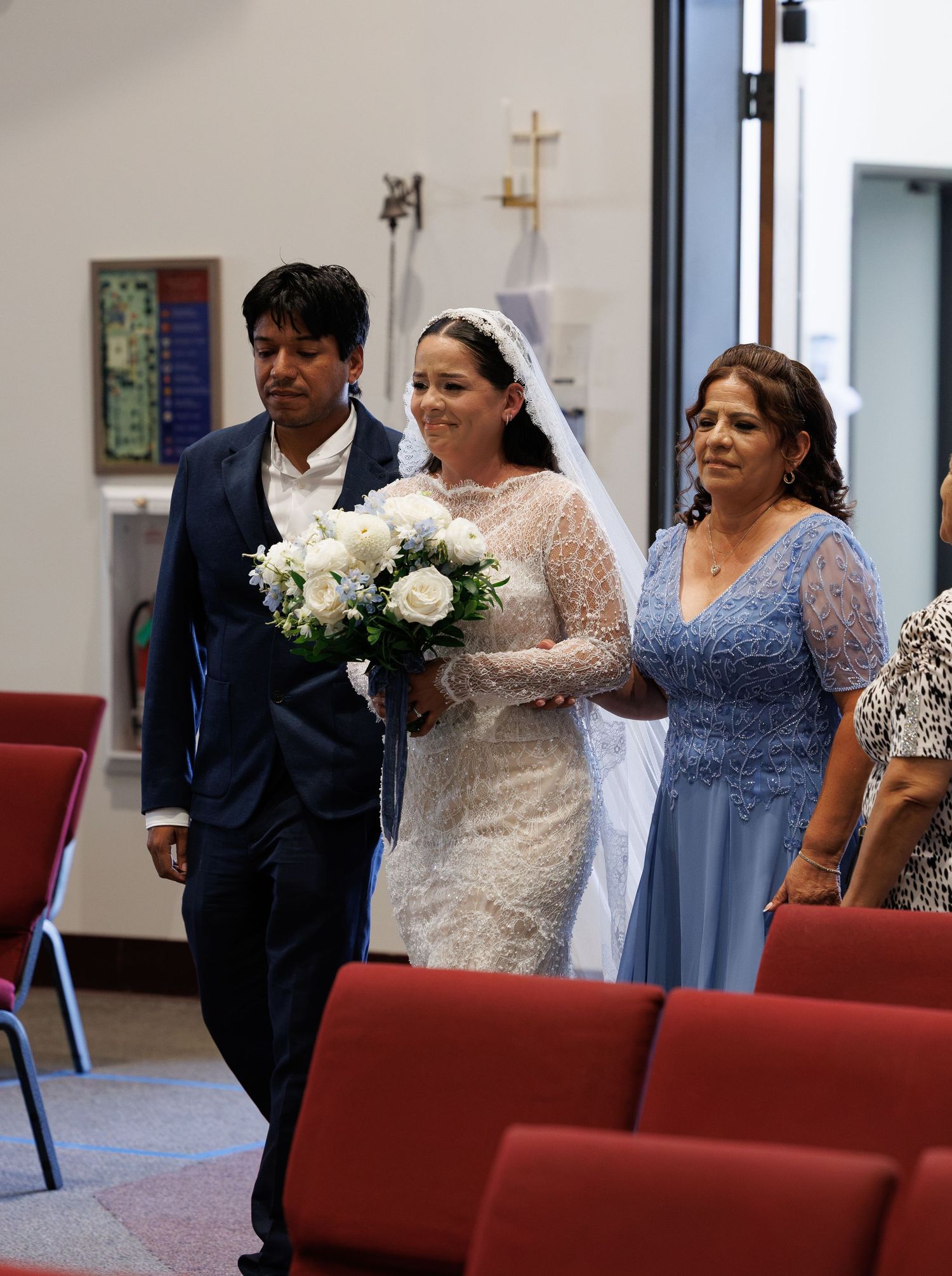 Bride in a lace gown and veil holding a white bouquet walks down the church aisle beside a man in a navy suit and a woman in blue.