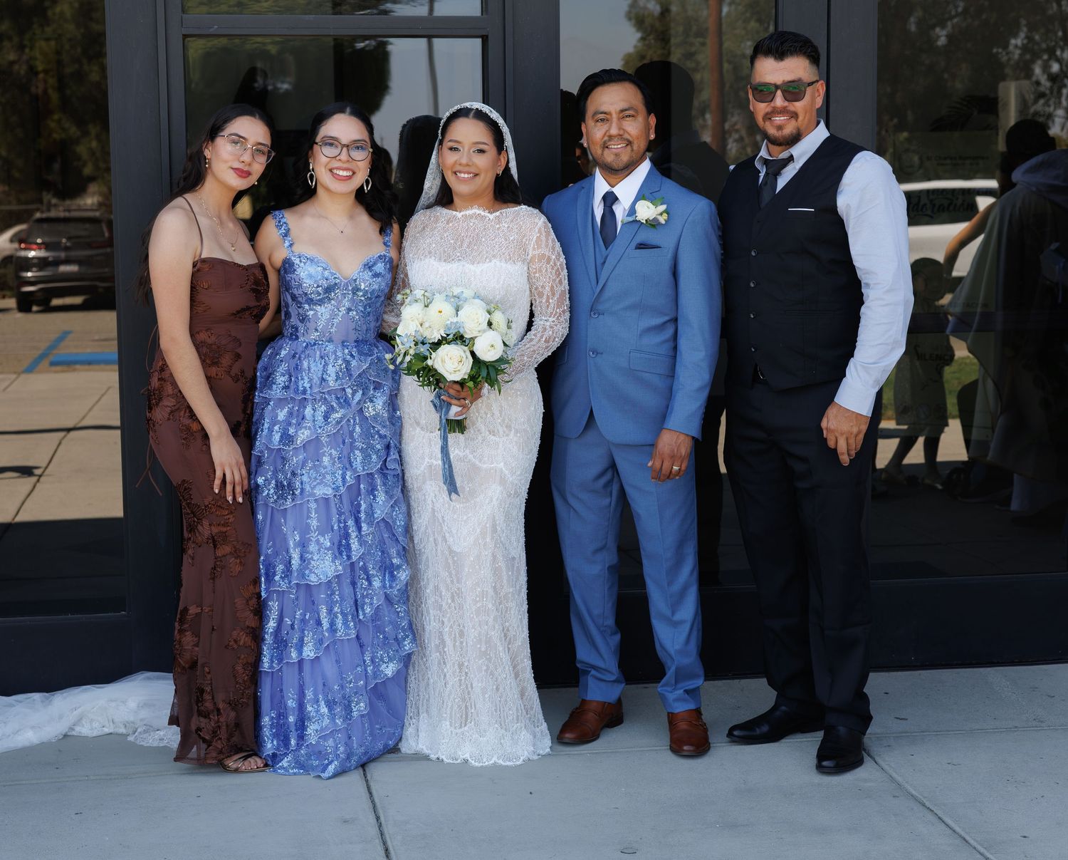 Bride in a white lace gown holding a white bouquet poses with a groom in a blue suit and three guests outside a glass building.