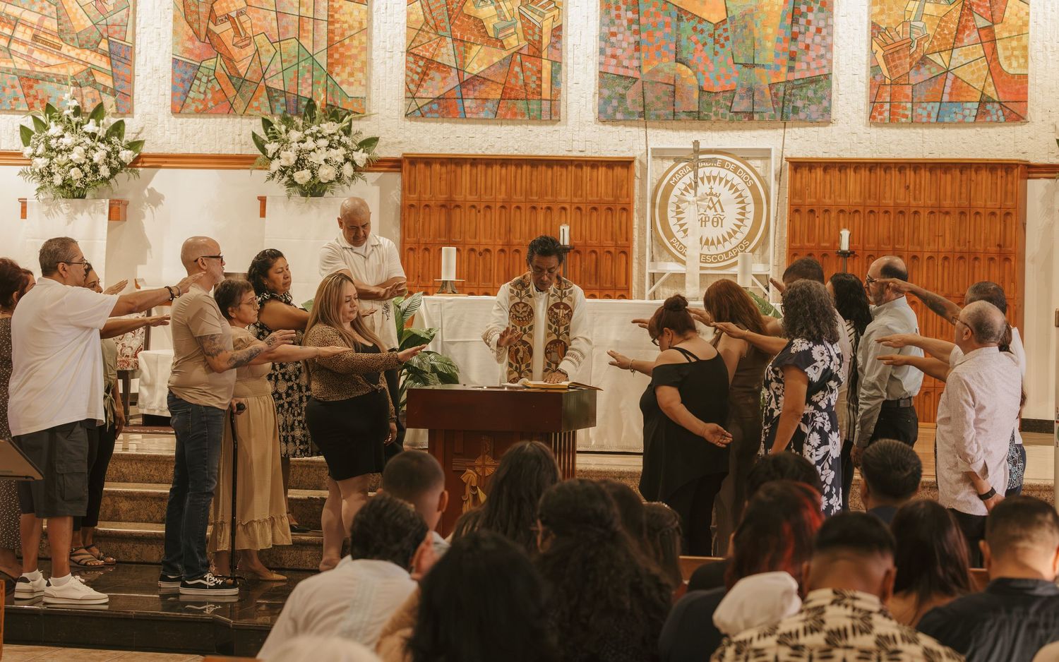 People gathered in a church ceremony near an altar with floral arrangements and colorful stained glass windows.
