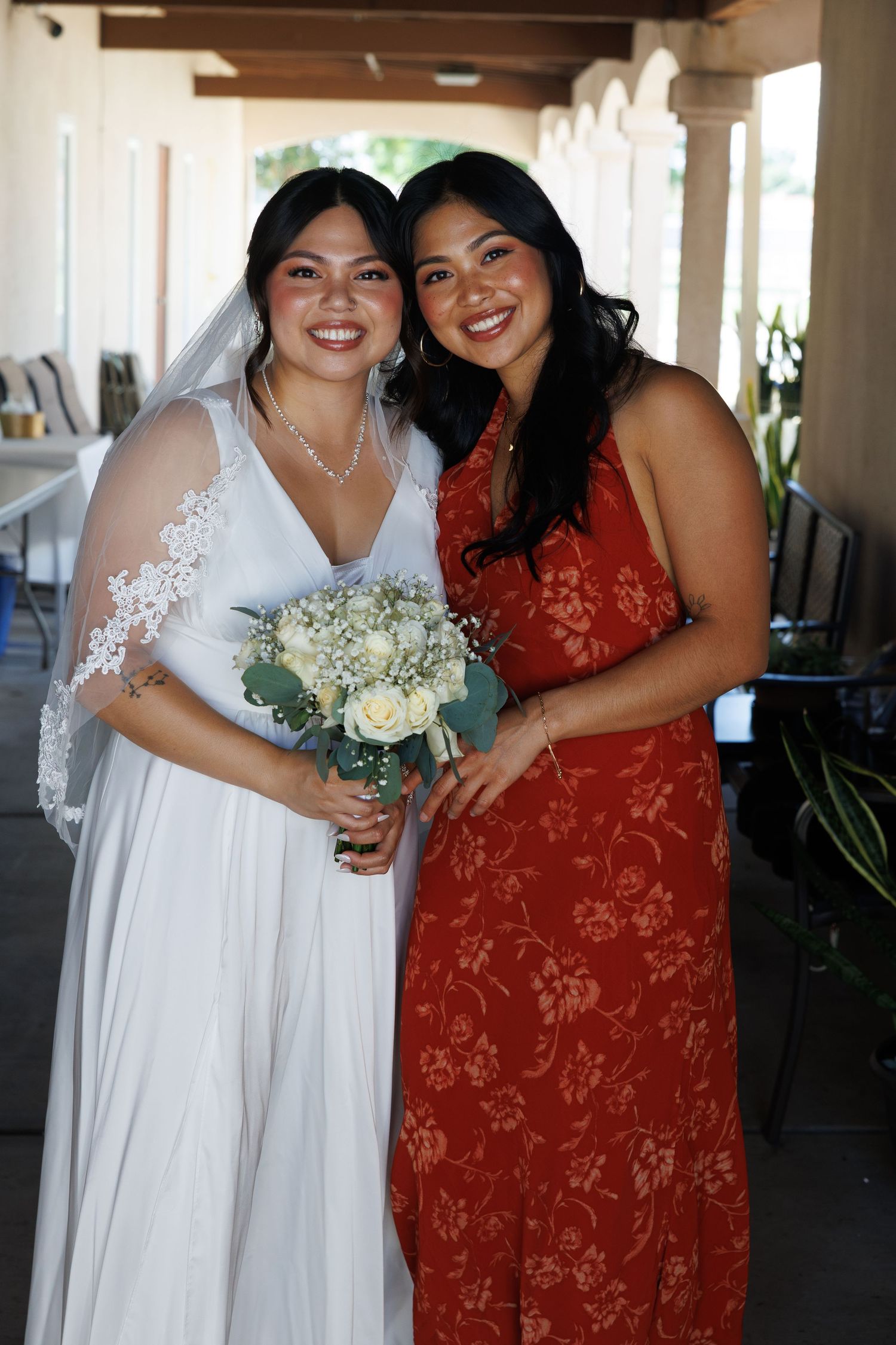 Bride in white lace veil holding bouquet poses with guest in red floral dress at outdoor wedding venue.