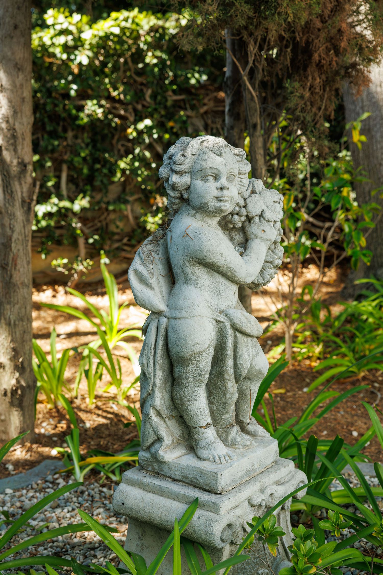 Stone cherub statue holding grapes surrounded by green foliage and trees in a sunlit garden.