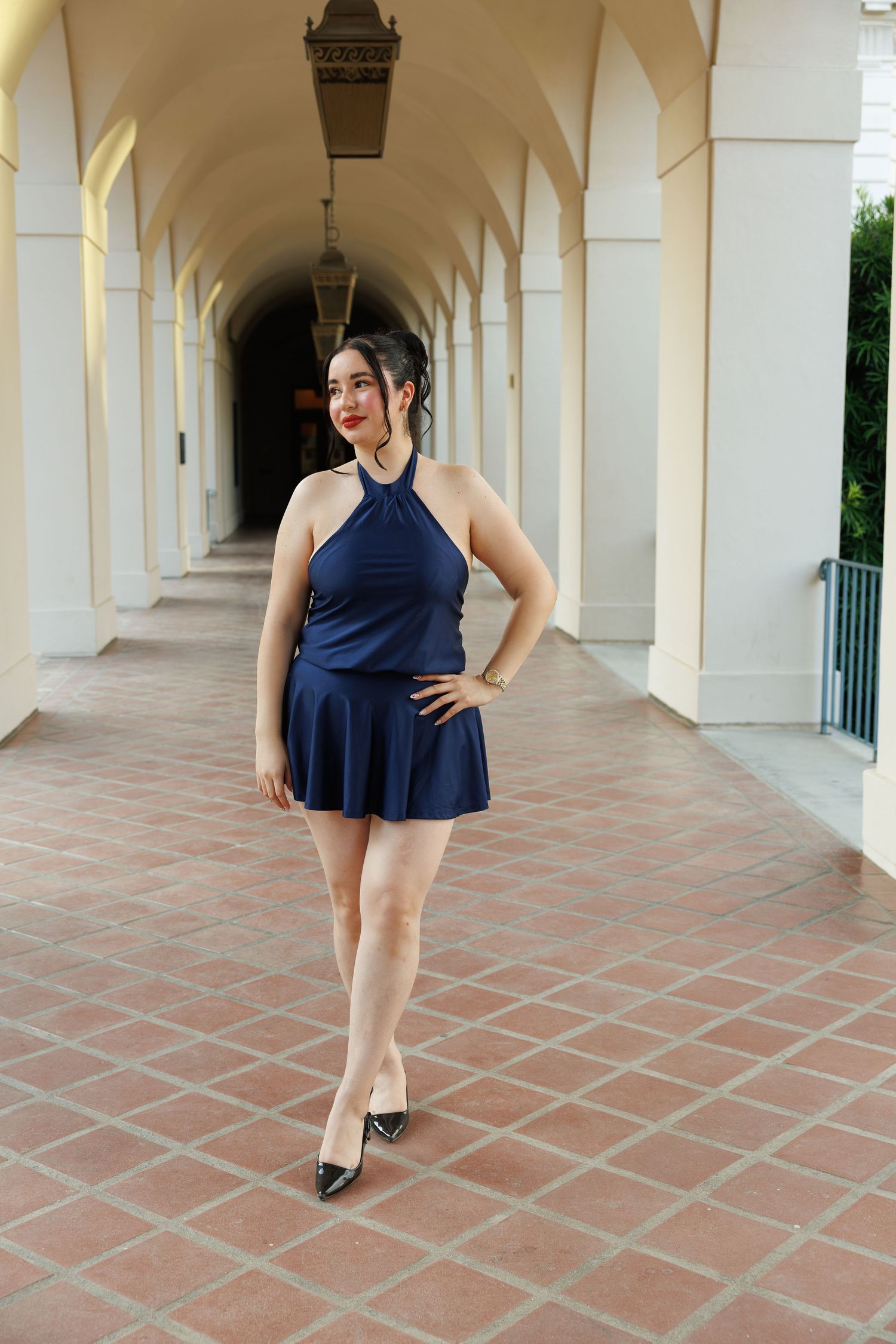 Woman in navy blue halter mini dress and black heels posing in elegant arched corridor with terracotta tile floor.