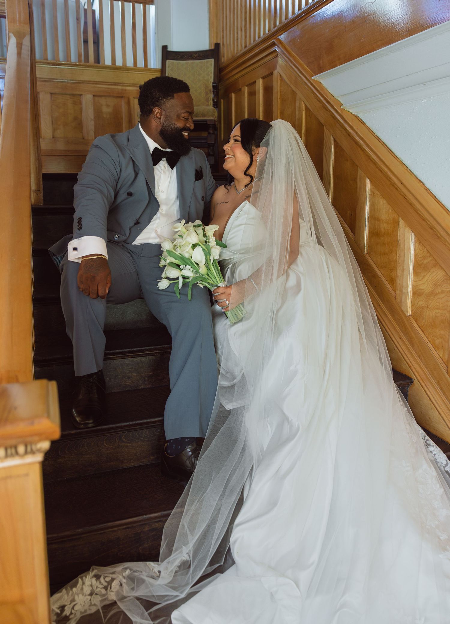 A bride in a white gown and groom in a gray suit share a romantic moment on wooden stairs.
